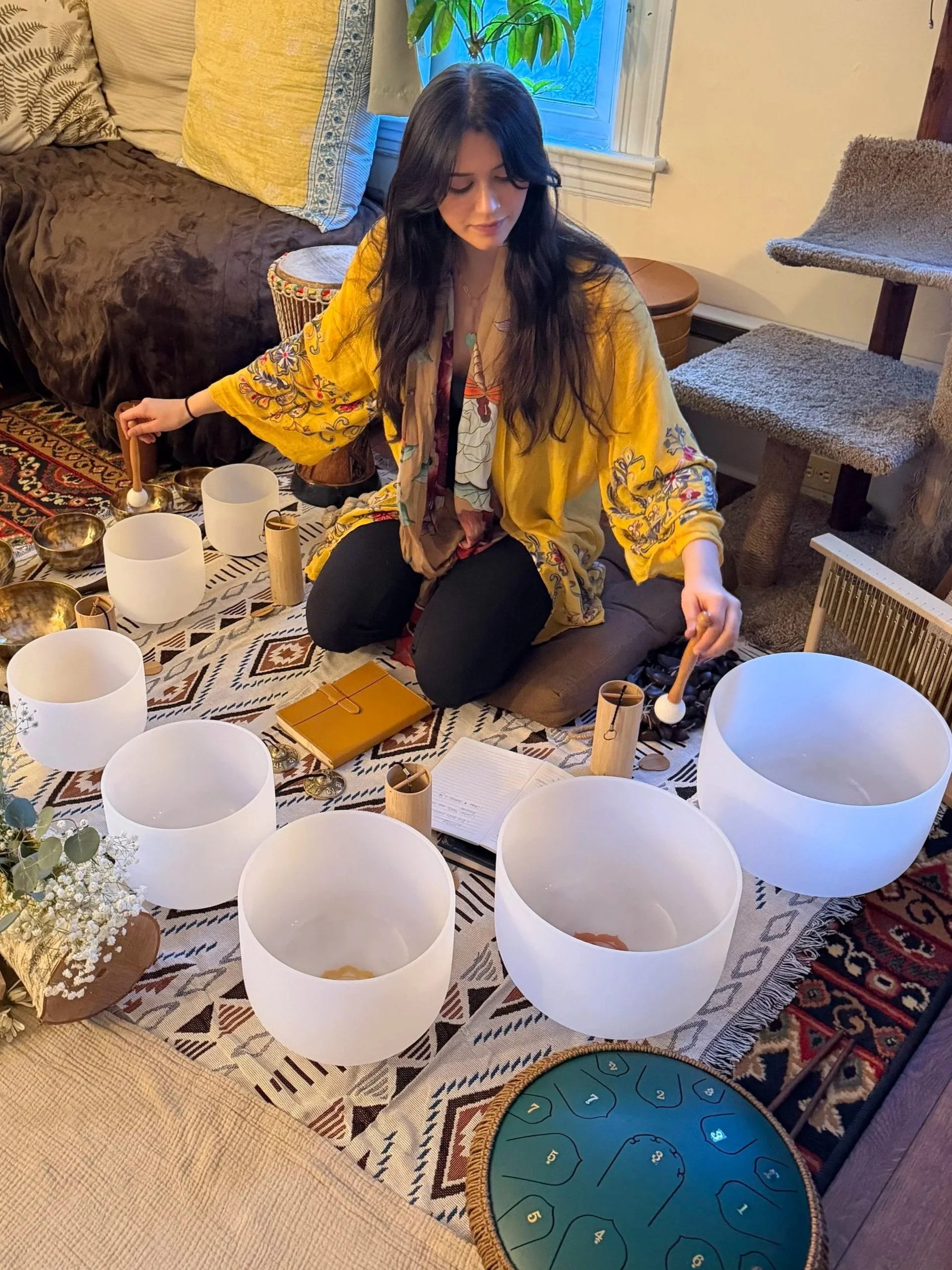 A woman is sitting on a patterned rug in a living room, playing crystal singing bowls with mallets. There are multiple bowls around her, a notebook, and a yellow wallet.