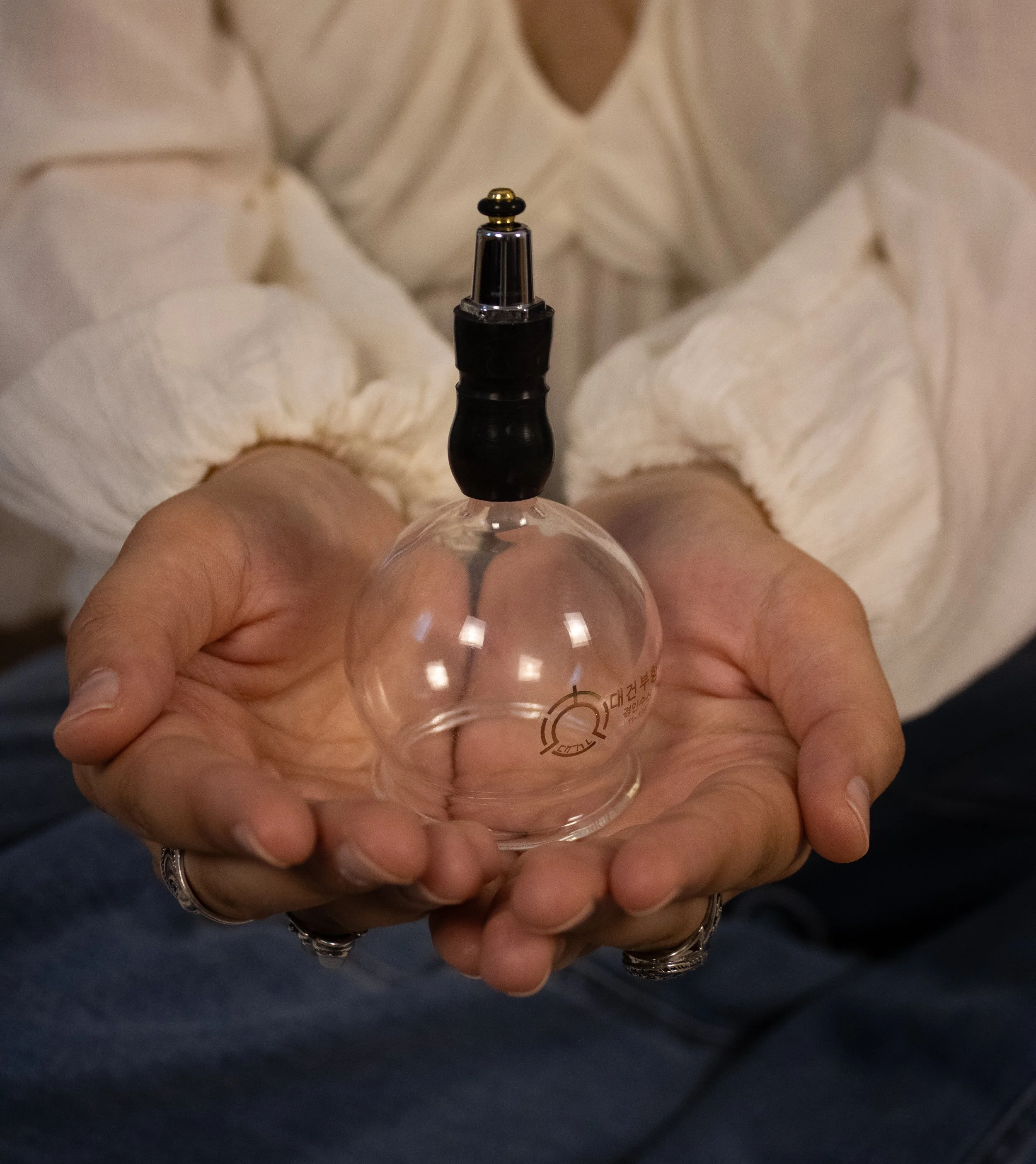 Close-up of a person's hand holding a clear glass spray bottle with a black and gold spray nozzle. The person is wearing a white and beige striped shirt and has jewelry on their fingers.