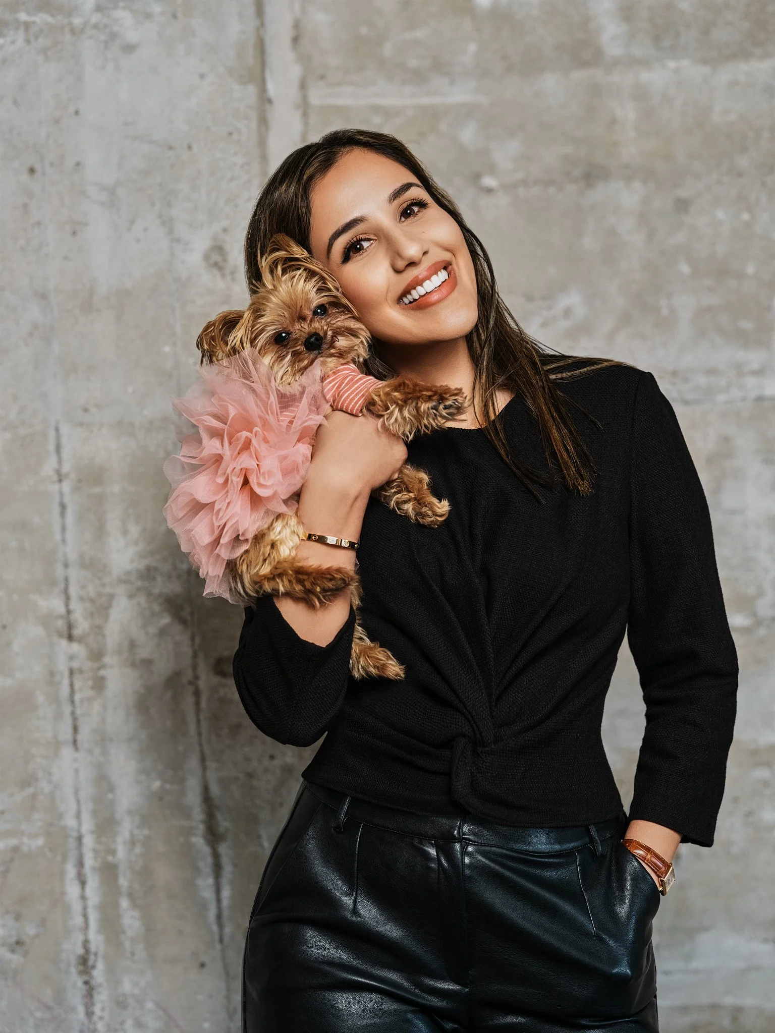 A smiling woman in a black long-sleeve top and black leather pants holding a small Yorkshire Terrier dog wearing a pink tutu and pink striped shirt.