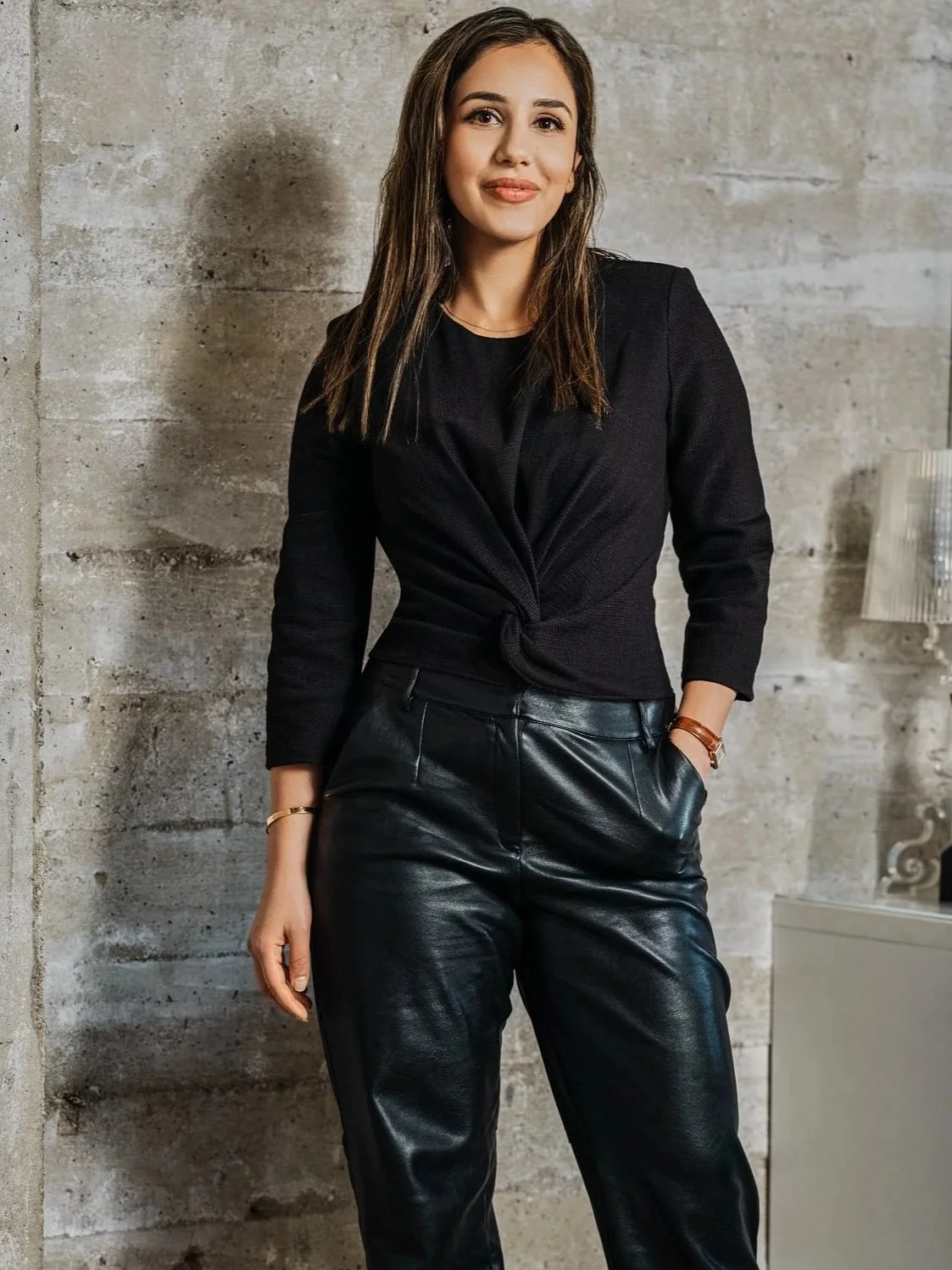 Woman standing in front of concrete wall, wearing a black top and leather pants.