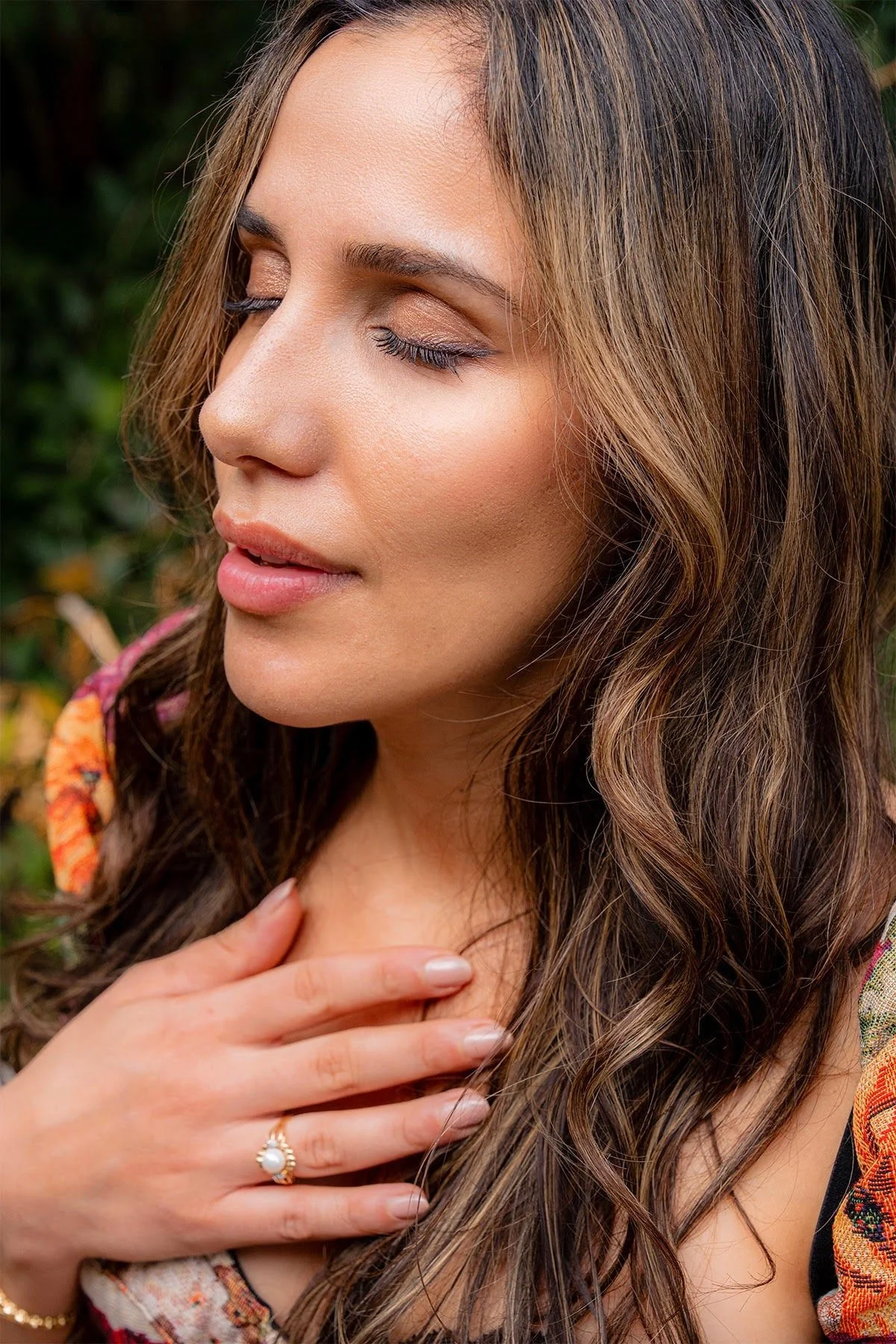 Close-up of a woman with long, wavy brown hair, closed eyes, and a relaxed expression, outdoors with greenery in the background.