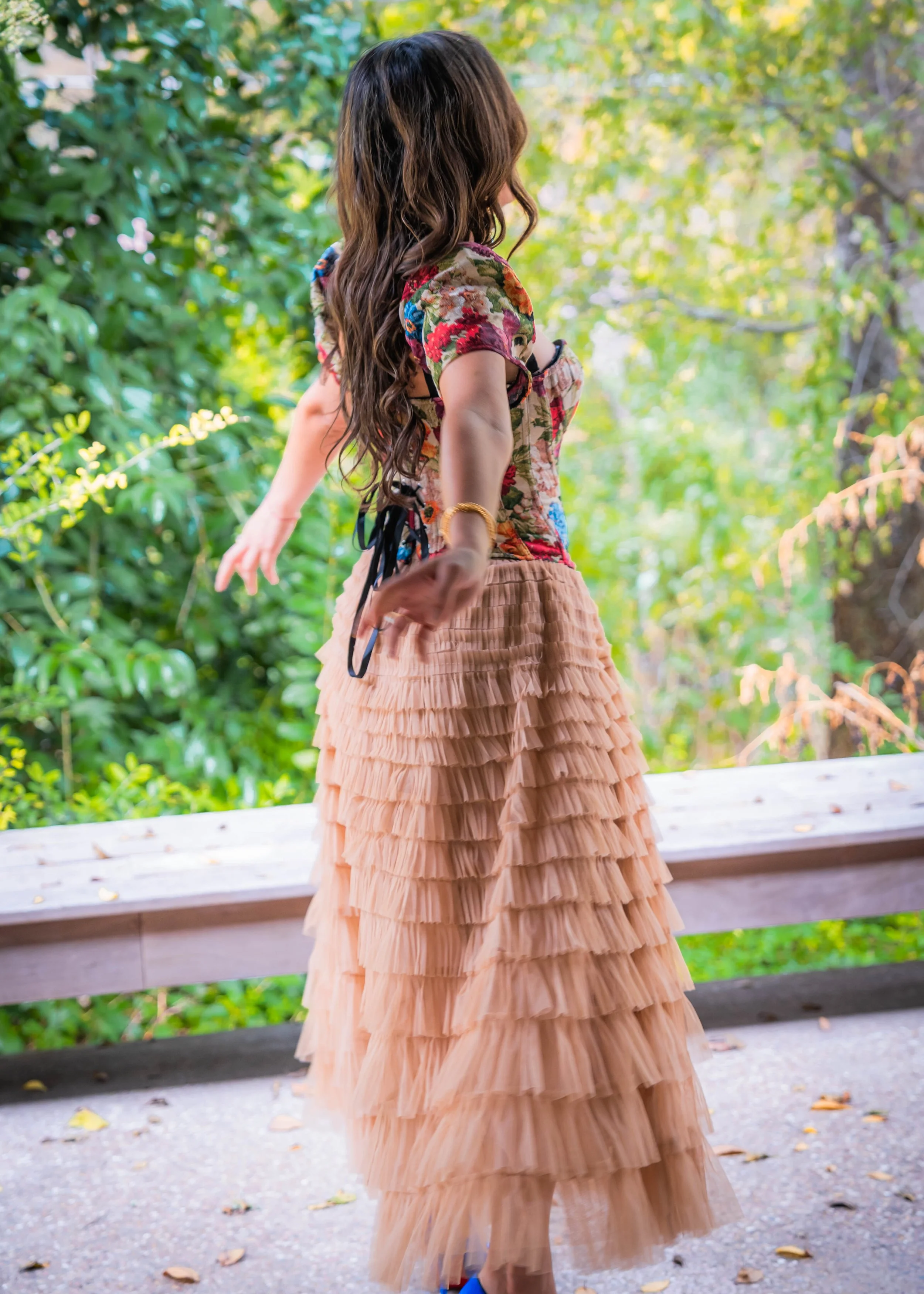 A woman with long brown hair, viewed from the side, balancing on a wooden bridge outdoors surrounded by greenery, wearing a colorful floral top and a long beige layered tulle skirt.