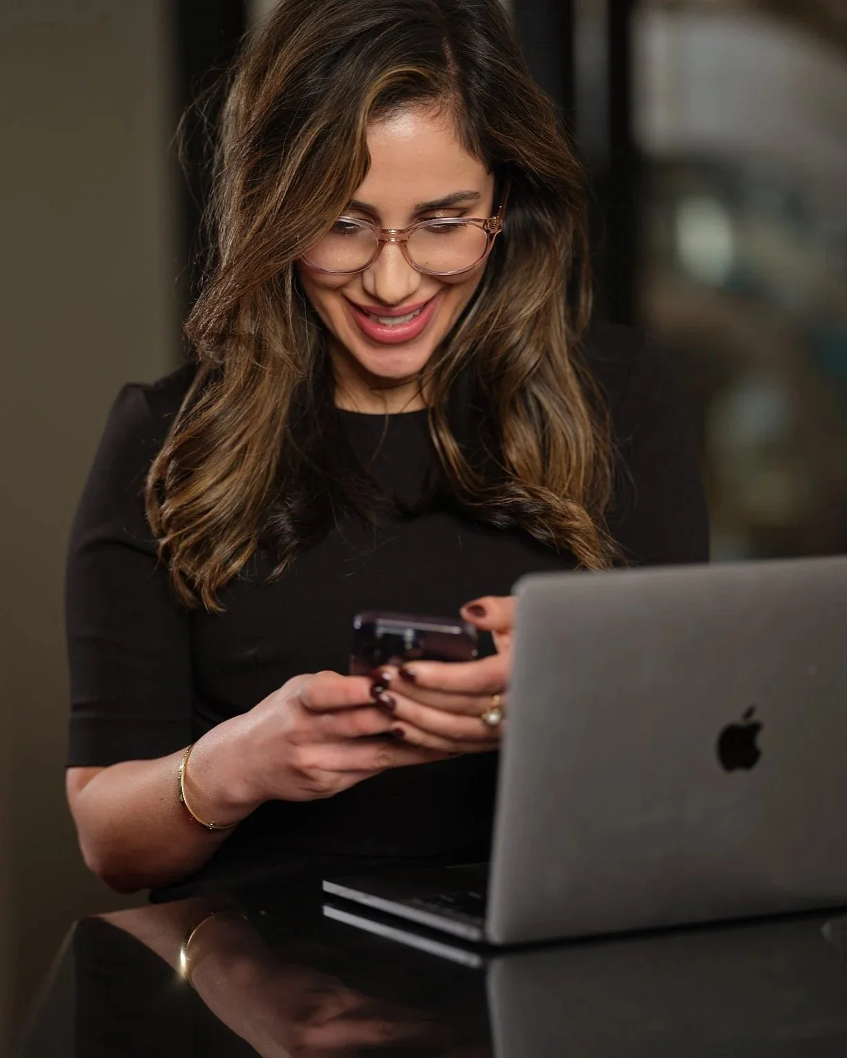 A woman with wavy brown hair, wearing glasses and a black shirt, sitting at a desk with a laptop, smiling and looking at her phone.