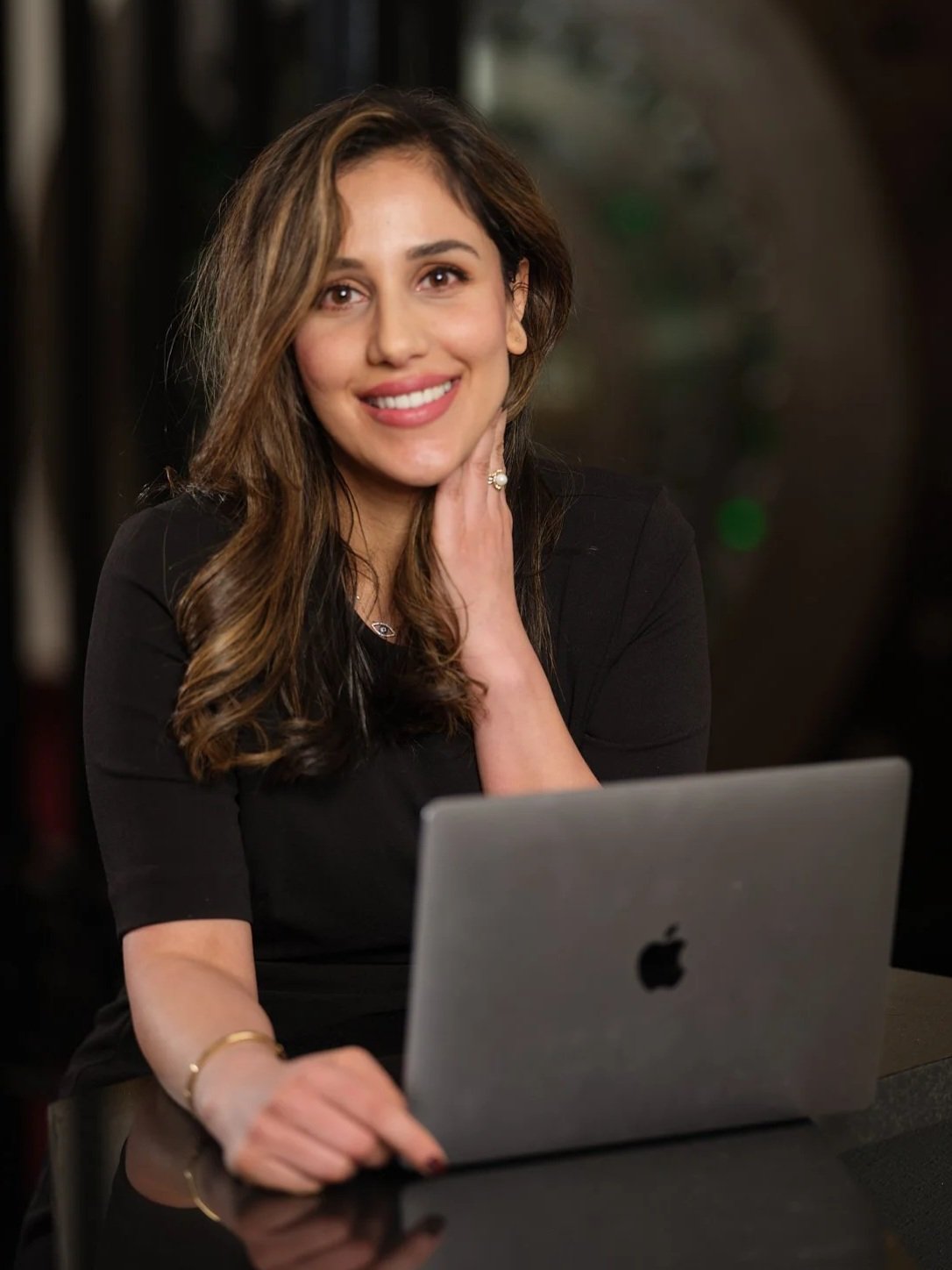 A woman smiling at the camera with hand resting on her neck, sitting at a table with a silver MacBook laptop in front of her.