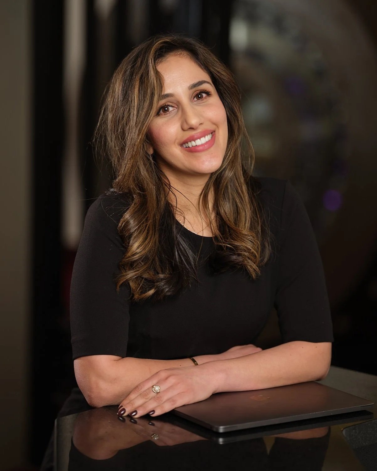 A woman with long brown hair, wearing a black top, smiling and sitting at a table with a closed laptop in front of her.