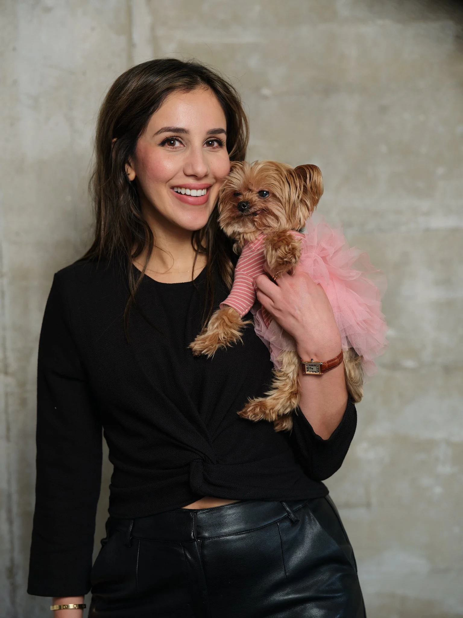 A woman with dark hair in a black long-sleeve top and black leather skirt holding a small dog dressed in a pink tutu and striped shirt, standing against a plain beige wall.