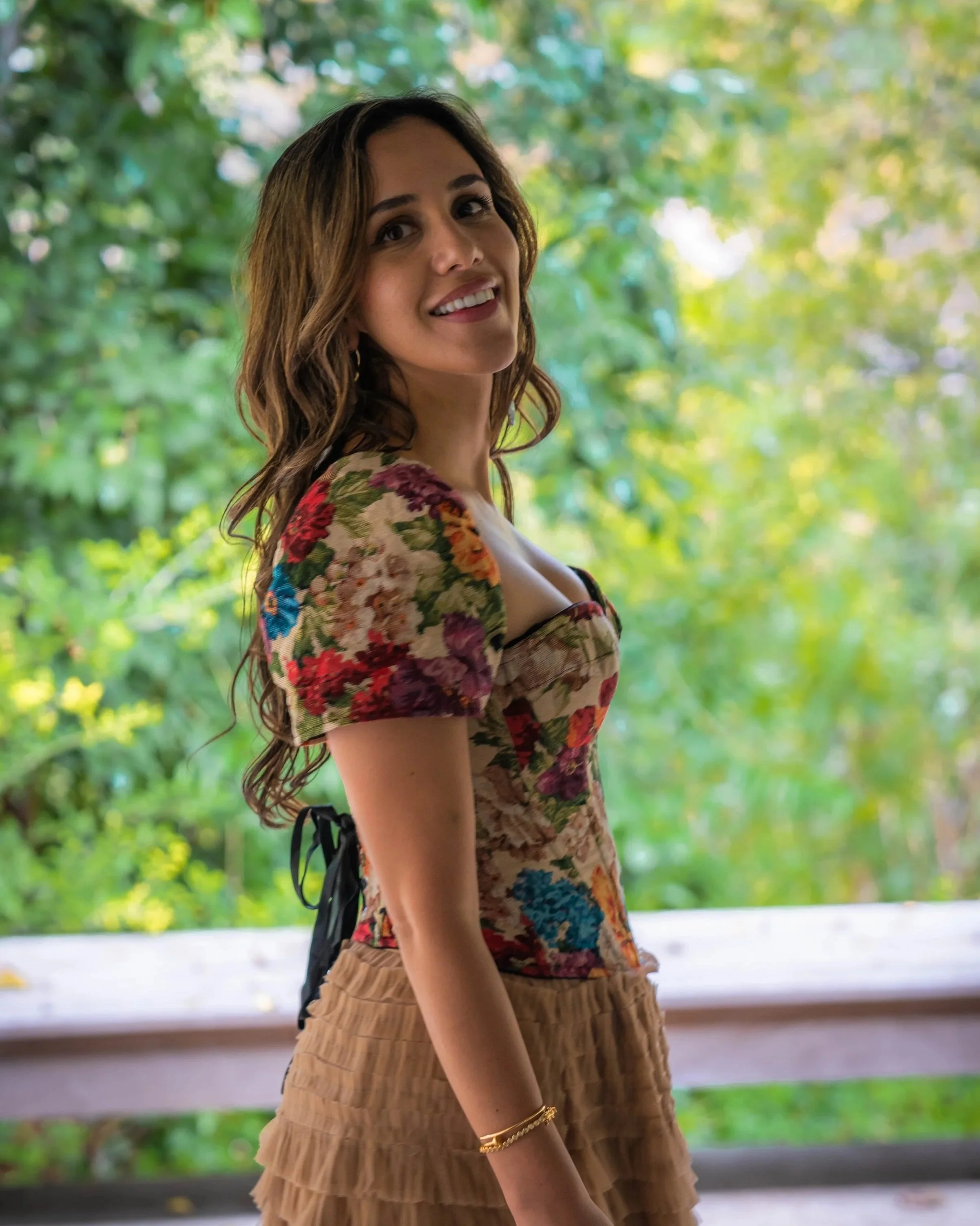 A woman with long wavy brown hair smiling outdoors with green trees in the background, wearing a floral top and ruffled beige skirt.