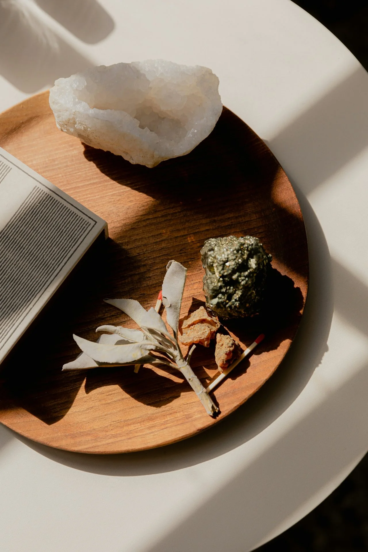 A round wooden tray holding a large white mineral, a dark metallic mineral, a dried white flower, and some small rocks. Part of a newspaper is visible at the edge of the tray.