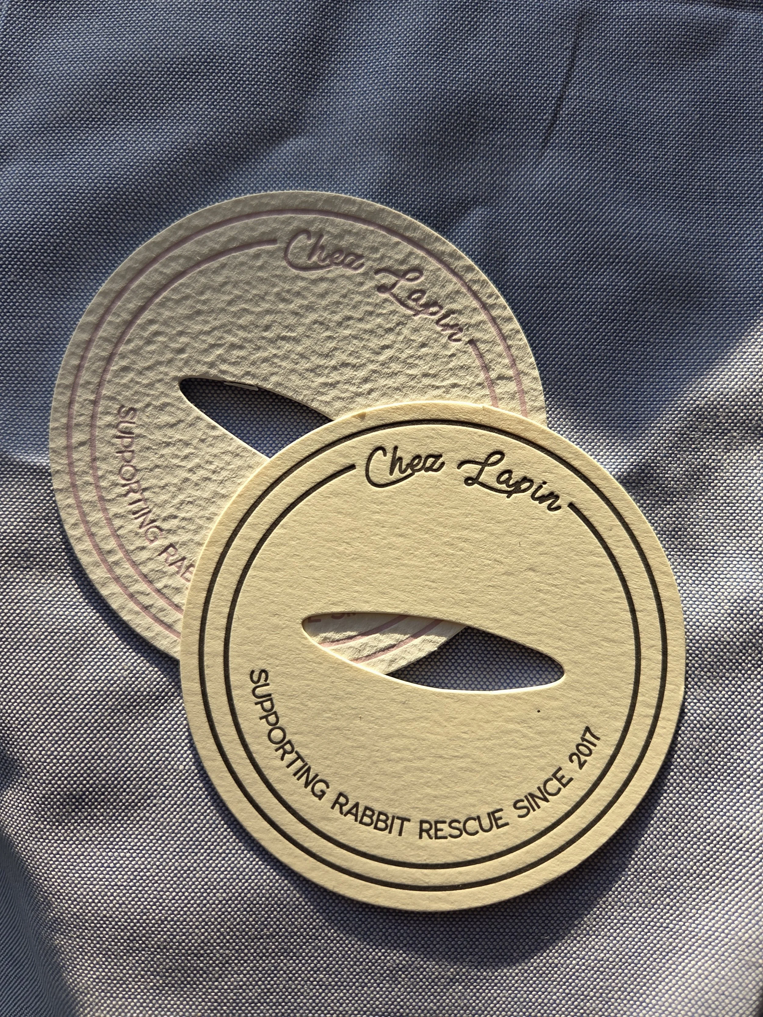 Two circular tokens with a slit in the middle and the words "Chez Lagin" and "Supporting Rabbit Rescue Since 2017" printed on them, placed on a textured fabric surface.