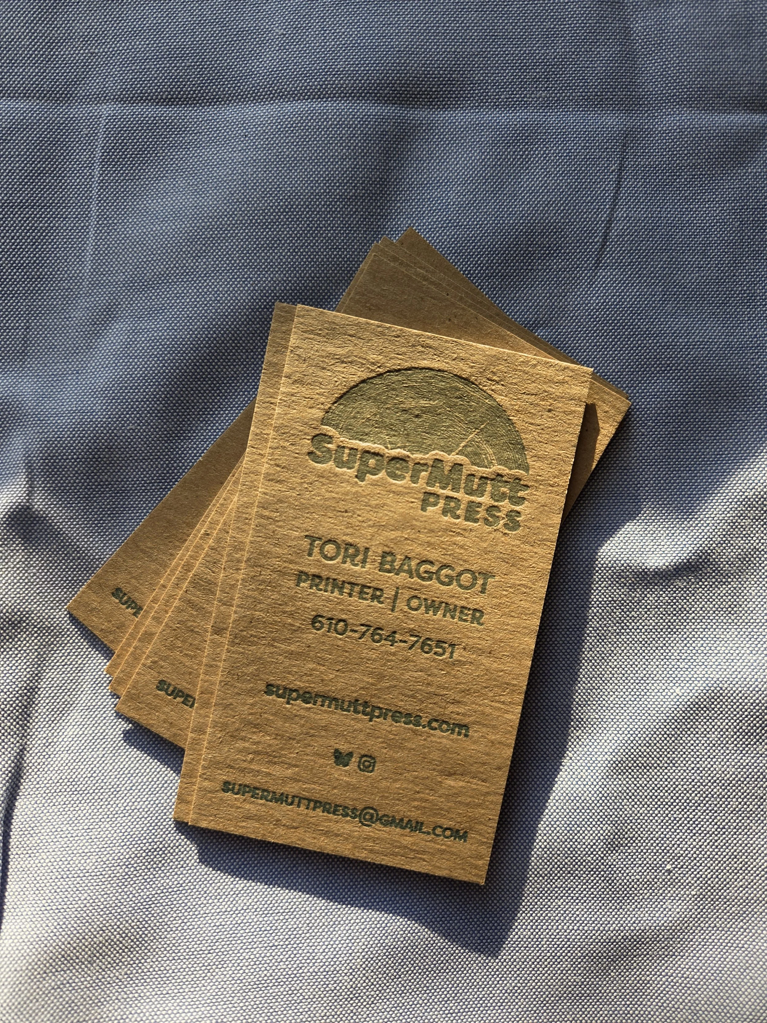 Business cards for SuperMutt Press, featuring contact information for Tori Baggot, a printer and owner. The cards are stacked on a textured light blue fabric background.