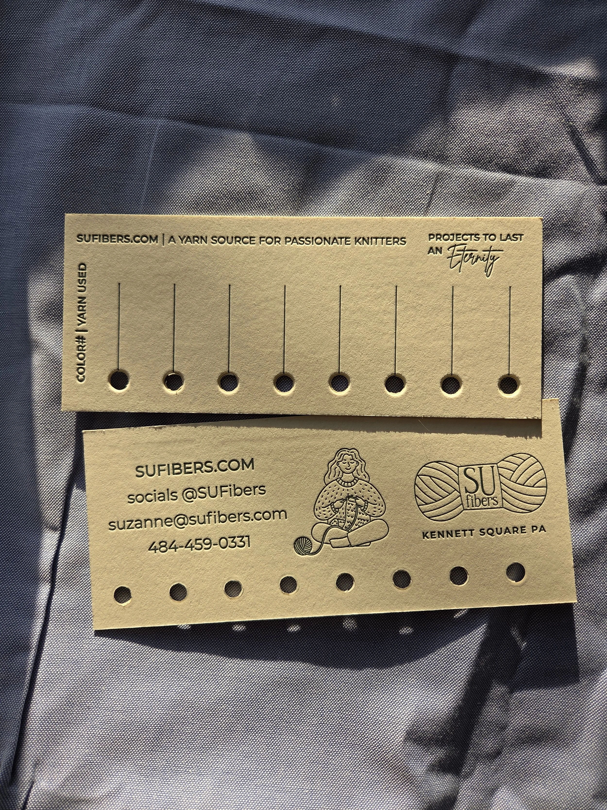 Business card and yarn sample card for SU Fibers, with contact info, logo, and a drawing of a woman knitting, placed on a fabric background.