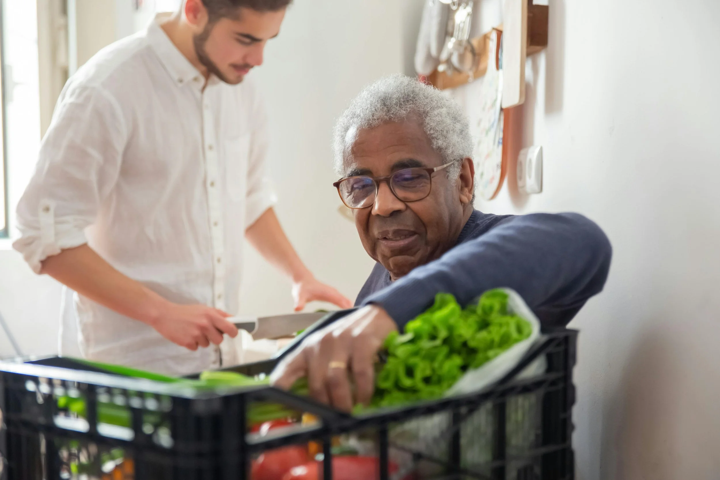 An elderly man with gray hair, glasses, and a black shirt reaches into a crate of fresh green lettuce, while a younger man in a white shirt stands beside him holding a knife, in a bright kitchen.