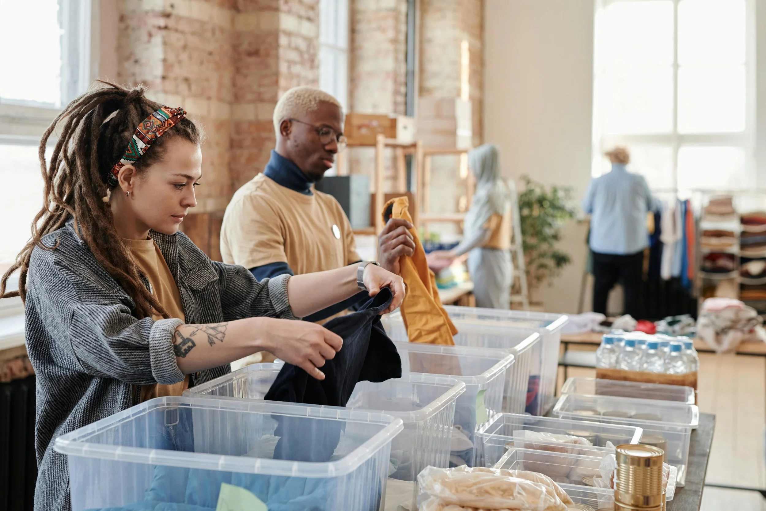 Two women sorting clothes into transparent plastic bins at a charity or thrift store. One woman has dreadlocks and tattoos, and the other has short blonde hair and glasses. In the background, two other people work near shelves of clothes and fabric.