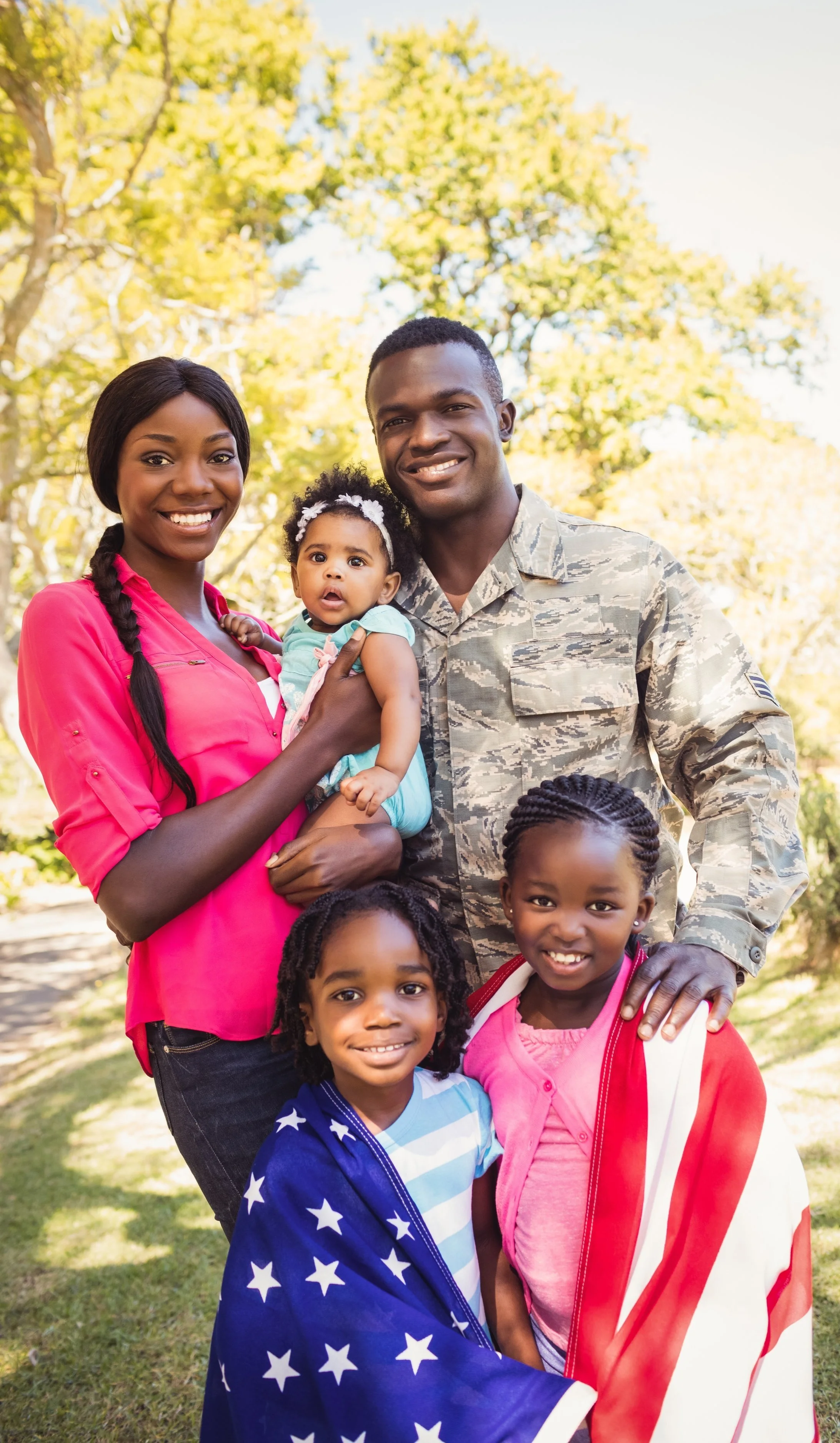 Family of six outdoors in a park with trees, with two adults, three children, and a baby, smiling for the camera, one child wrapped in an American flag.