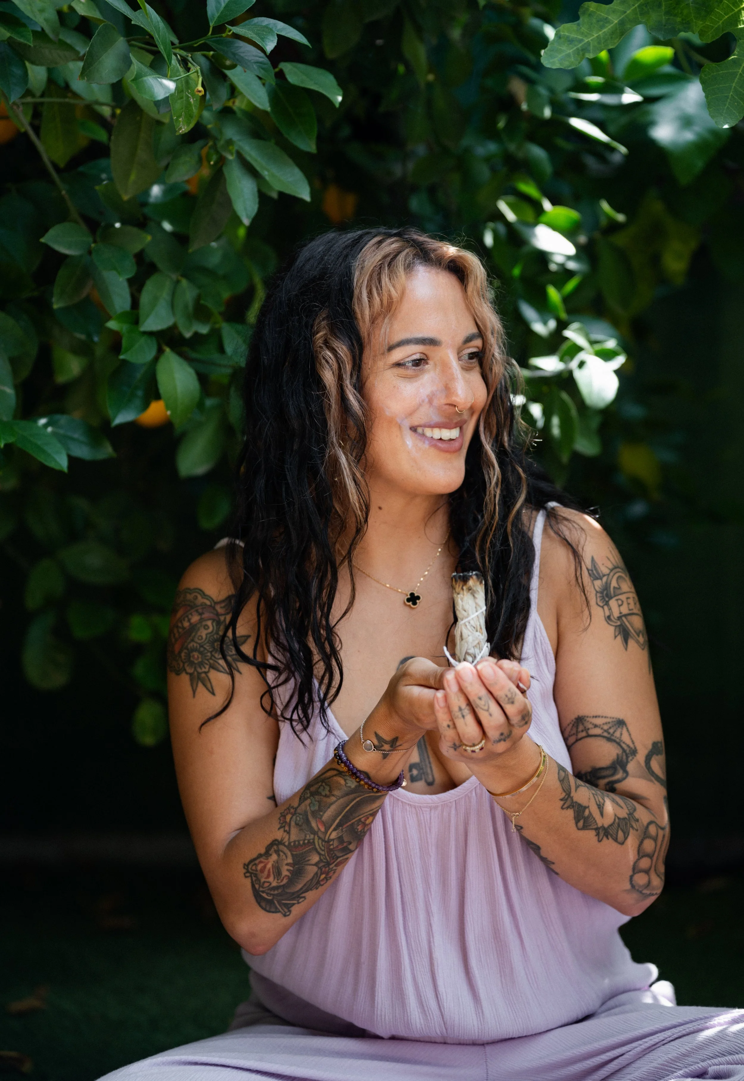 A woman with tattoos and wavy black and blonde hair smiling and holding a small bird in her hands outdoors, surrounded by green foliage