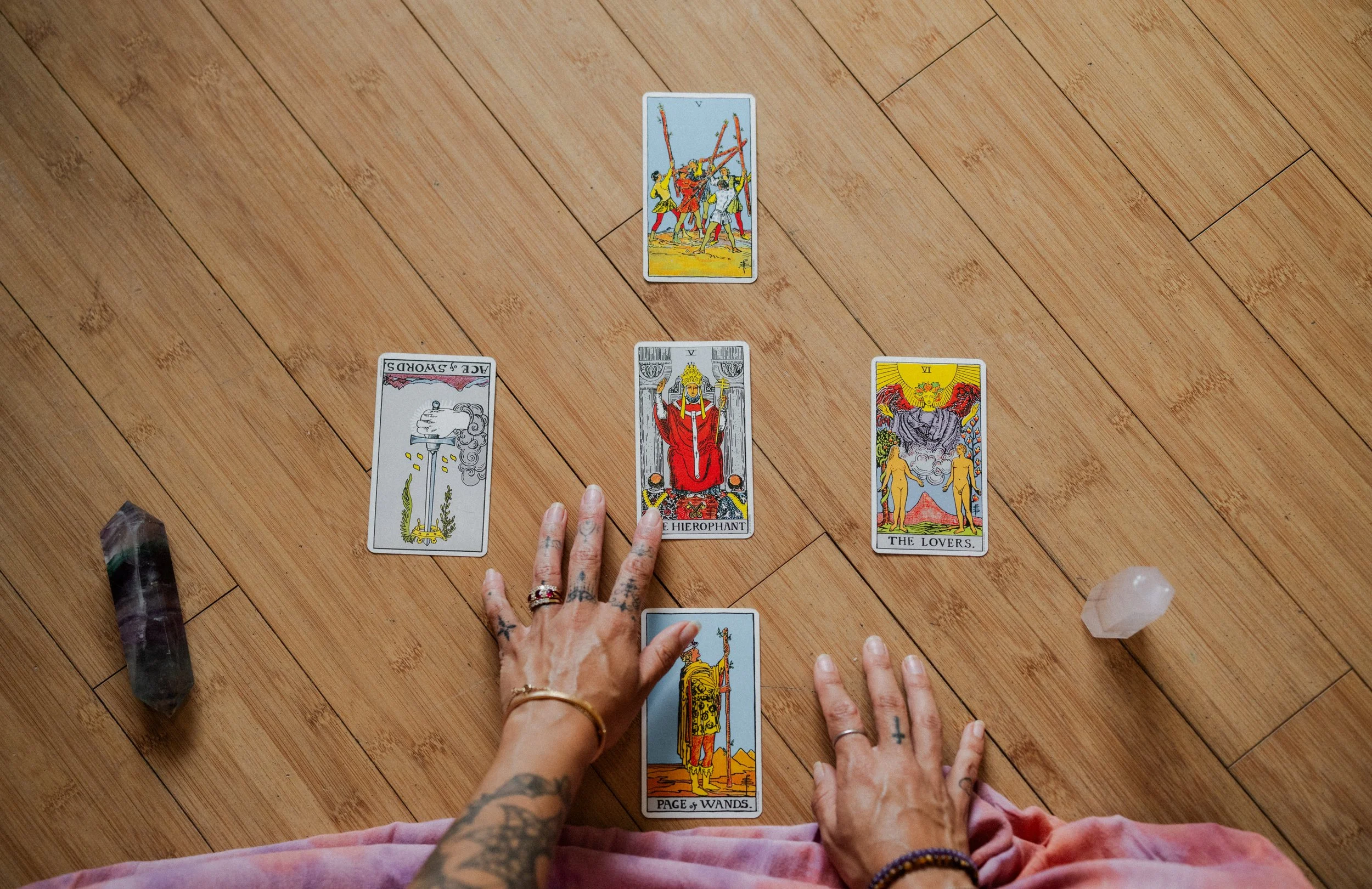 Person with tattoos and rings arranging tarot cards on a wooden floor, with a crystal and a stone nearby.