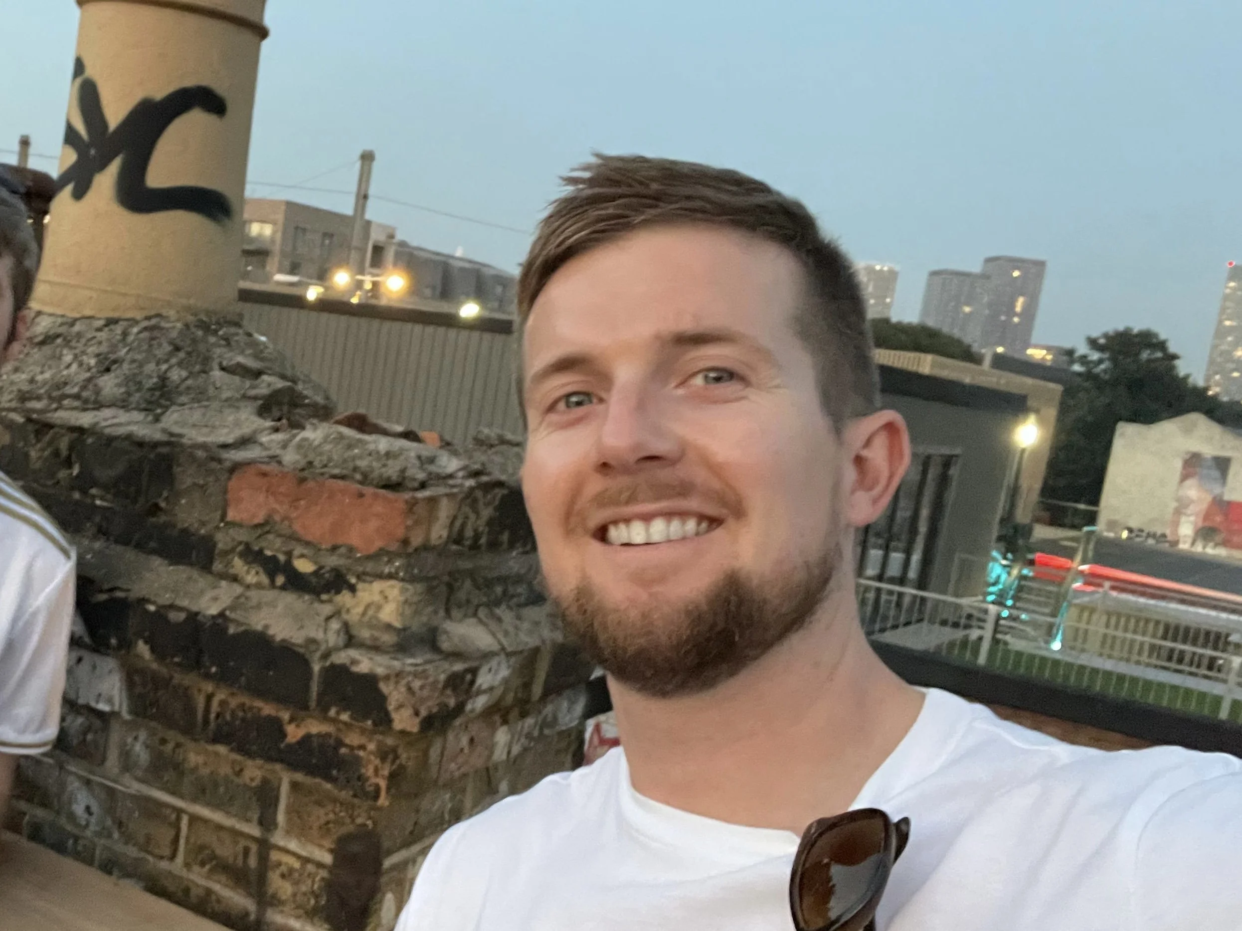 A smiling man with short brown hair, a beard, and wearing a white t-shirt taking a selfie outdoors. In the background, there is a brick and stone structure, a wooden pole with black graffiti, and city buildings with lit windows at dusk.