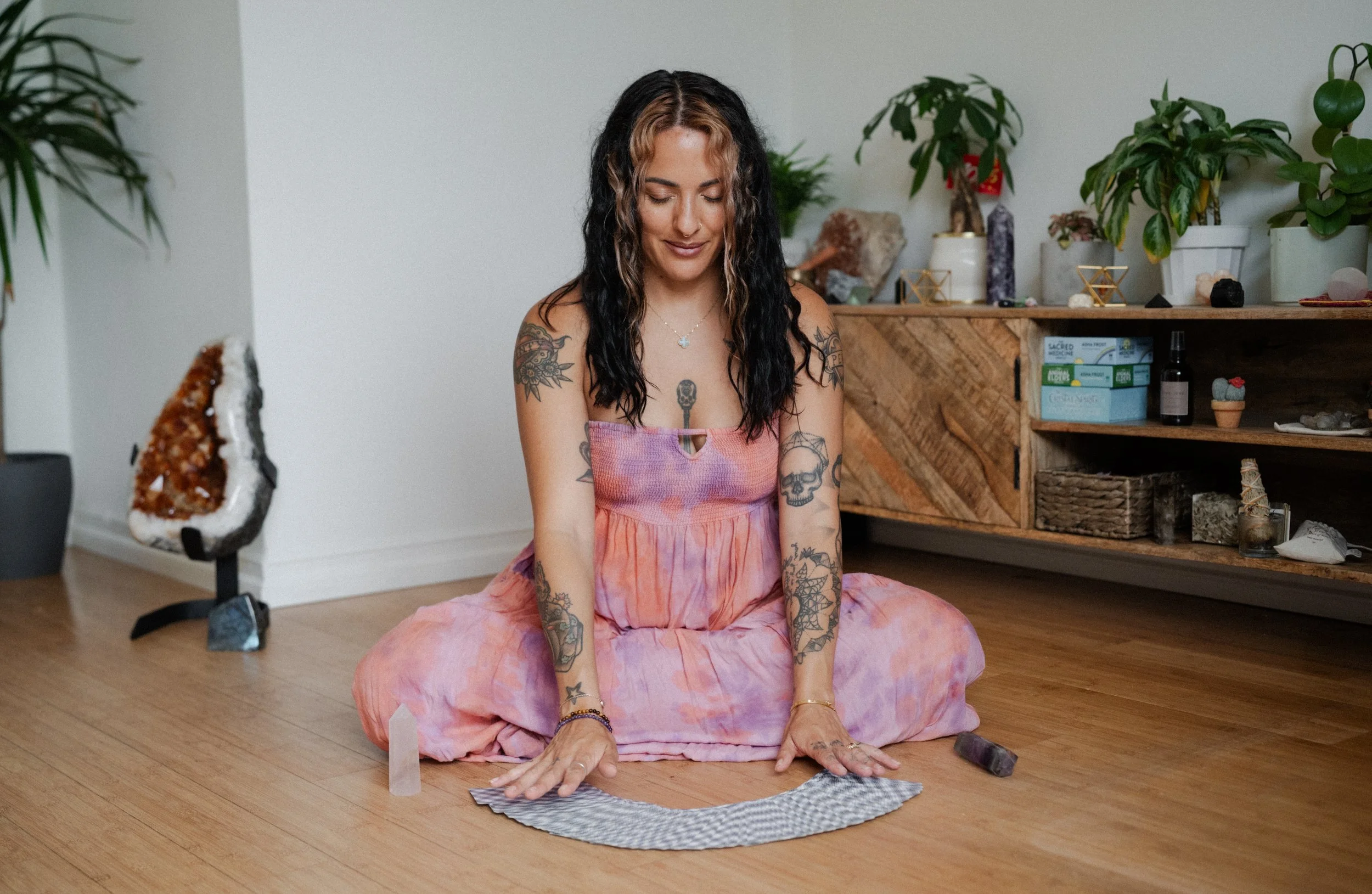 A woman with tattoos sitting cross-legged on the floor in a room decorated with plants and crystals, with a large crystal on a stand nearby, and a wooden cabinet against the wall. Doing a Tarot reading.