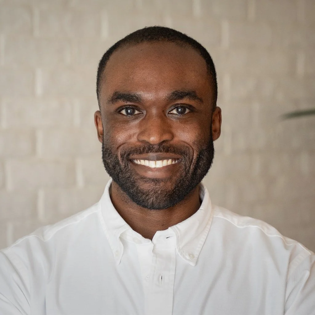 Headshot of a smiling African British man with short hair and a beard, wearing a white button-up shirt, standing in front of a light-colored brick wall.