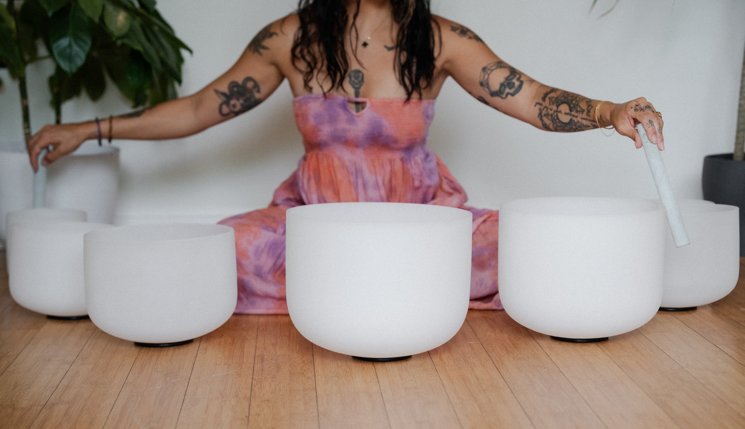 Person sitting on the floor playing white crystal singing bowls, wearing a colorful tie-dye dress, with tattoos on their arms and dark curly hair, indoors with a houseplant in the background.