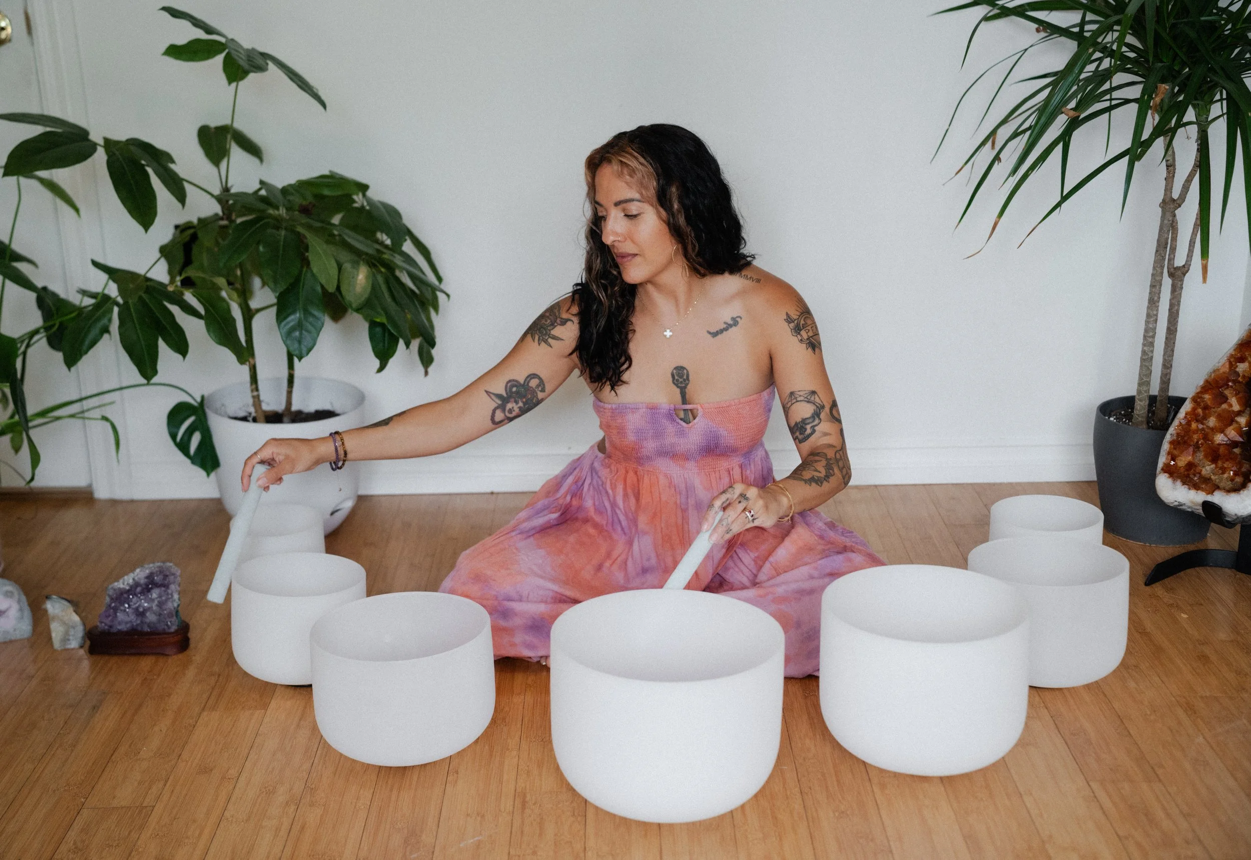 A woman with tattoos wearing a colorful strapless dress sitting cross-legged on a wooden floor, playing white crystal singing bowls surrounded by plants and crystals.