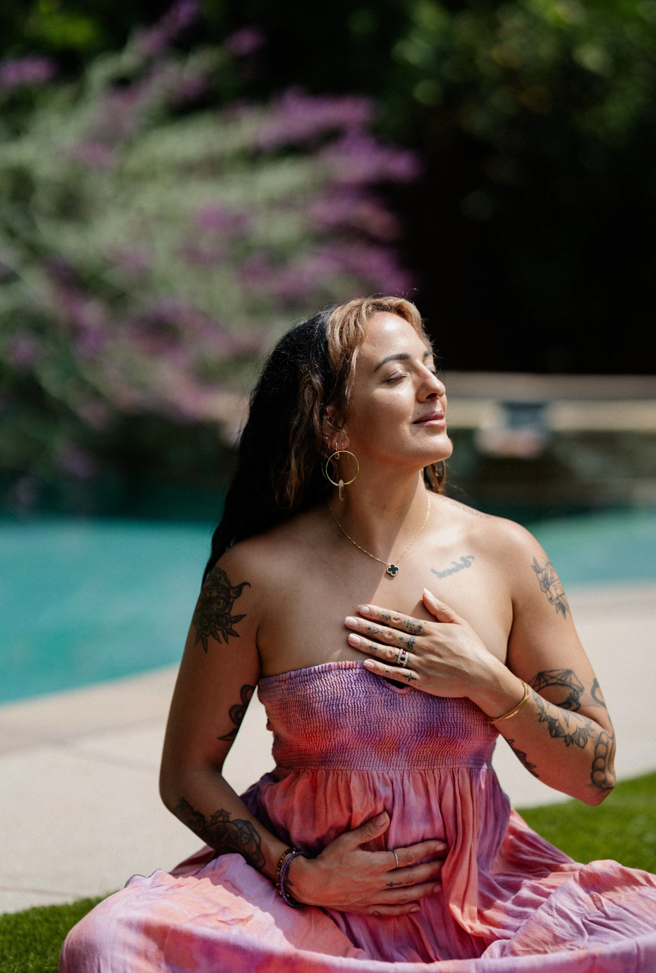A woman with tattoos sitting cross-legged outdoors near a swimming pool, eyes closed, in a peaceful pose with one hand on her chest and another on her belly, wearing a strapless pink dress and jewelry.