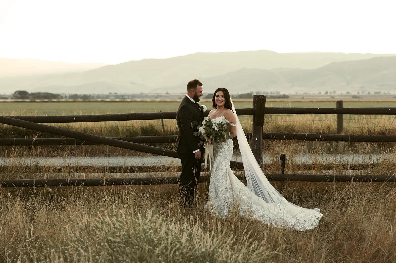 Bride and groom standing near a wooden fence in a field, with mountains in the background. The bride is in a white gown holding a bouquet, and the groom is in a dark suit.