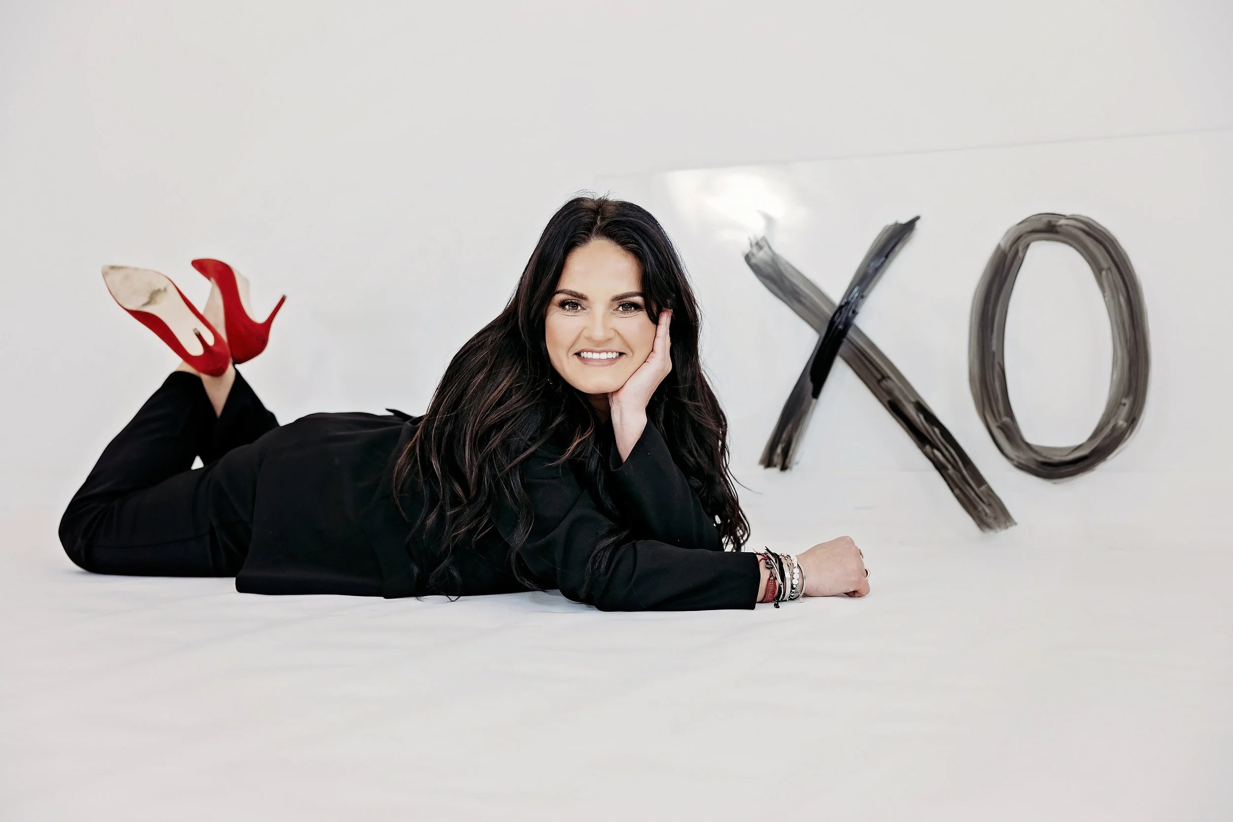 Woman in black outfit and red heels laying on floor with "XO" art in background.