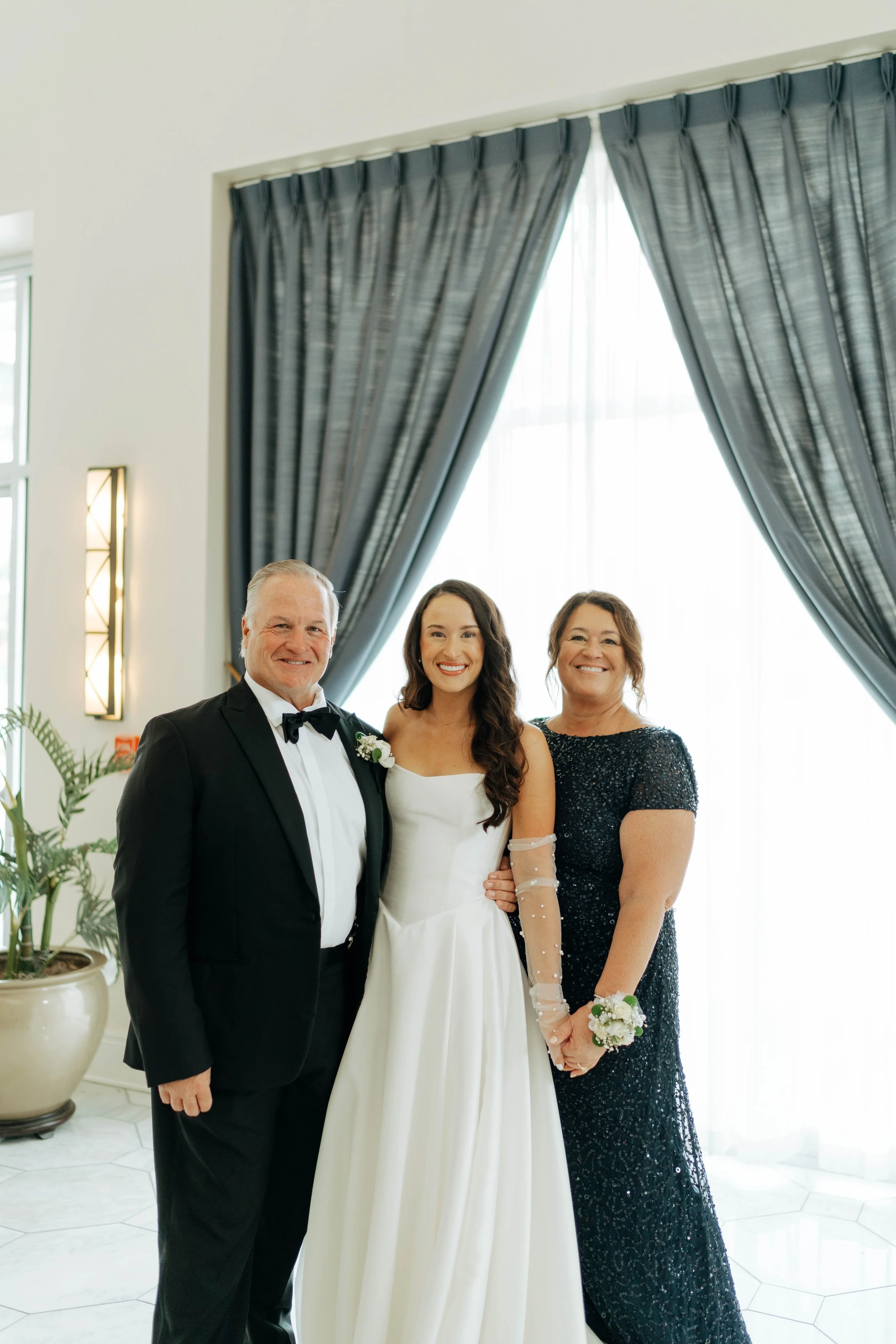 A bride in a white wedding dress stands between two elegantly dressed adults in formal attire, posing happily indoors in front of a window with dark curtains.