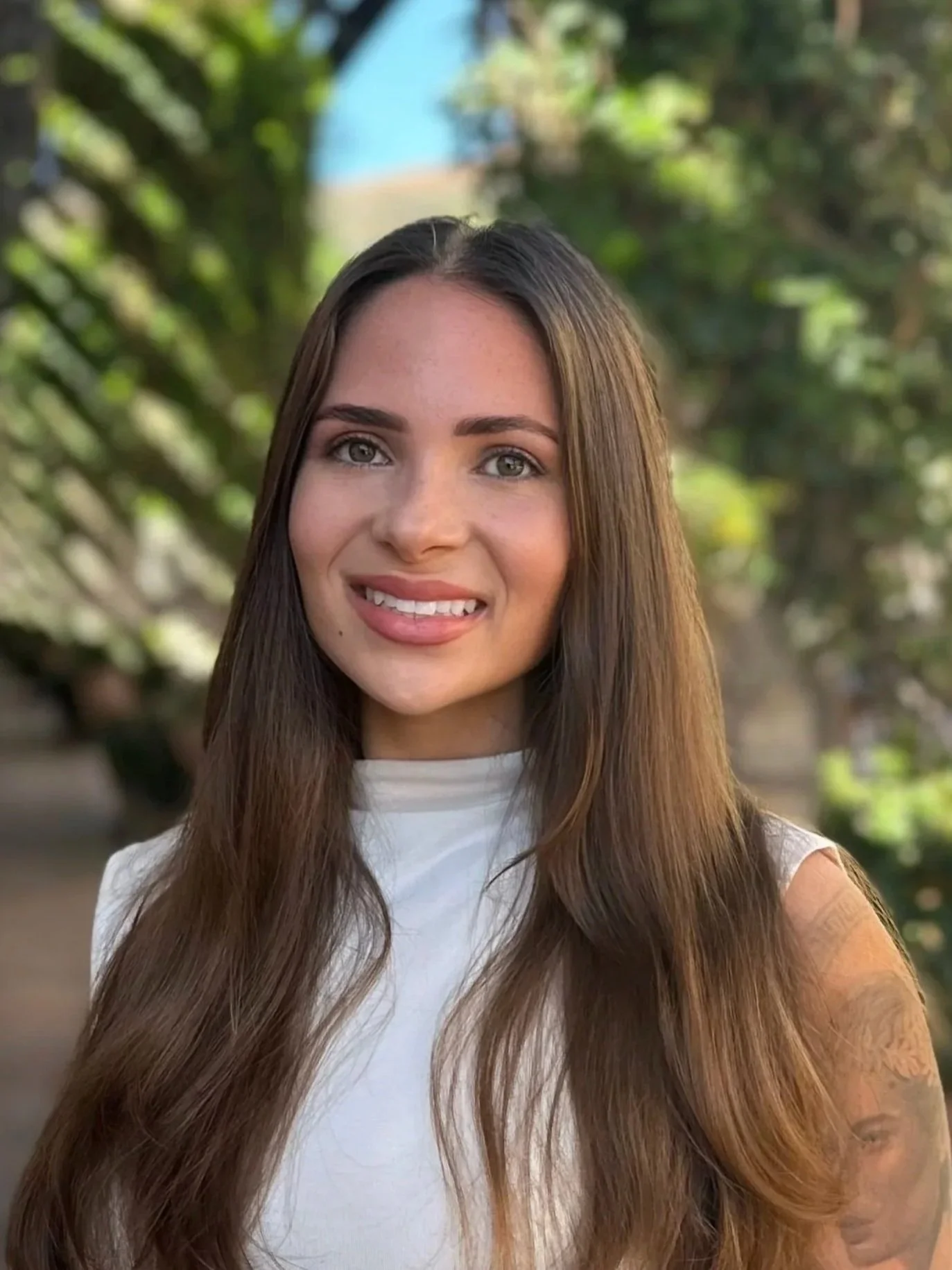 A young woman with long brown hair and light skin smiling outdoors with trees in the background.