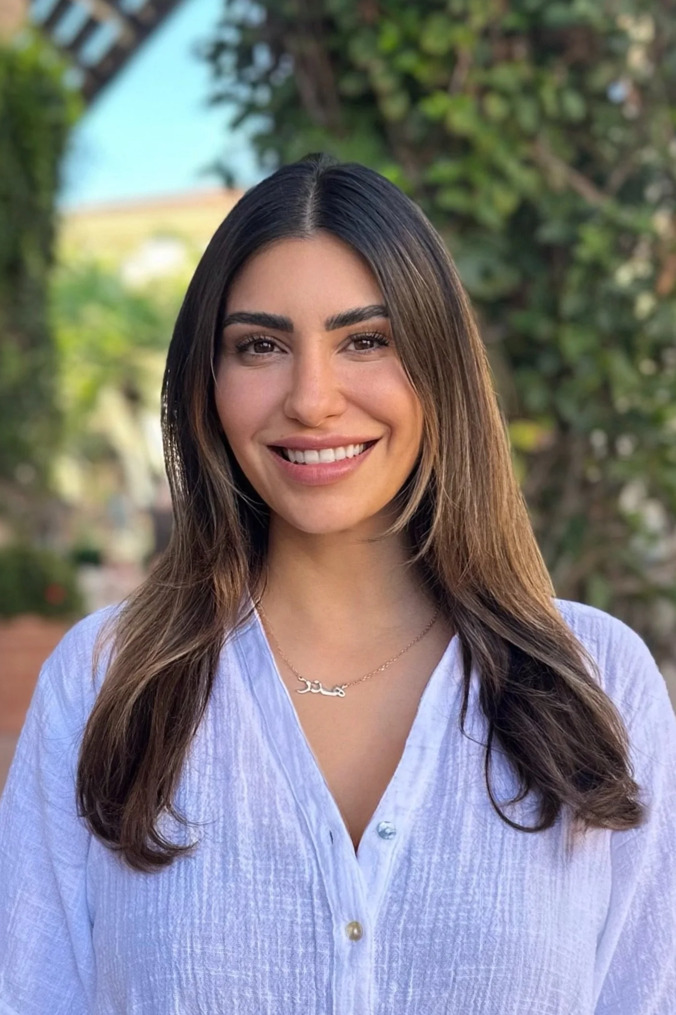 Headshot of a young woman with long wavy brown hair, wearing a light blue button-up shirt and a gold necklace, standing outdoors with greenery and trees in the background, smiling.