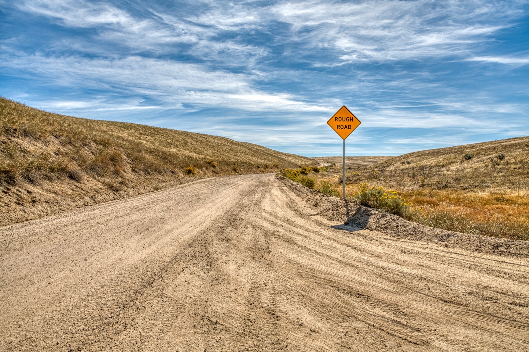 A winding dirt road leads through the golden Idaho foothills under a wide, cloud-brushed sky in this rural landscape photograph. The Rough Road sign hints at adventure ahead, inviting travelers to slow down and embrace the rugged beauty of open count