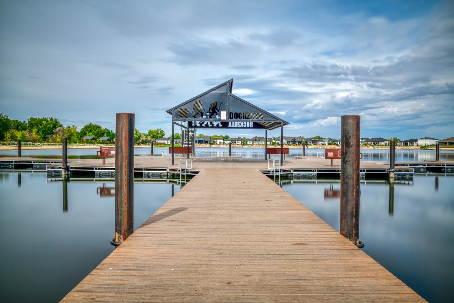 A smooth, glass-like surface transforms the lake at Freedom Park into a mirror in this long-exposure photograph of the Doczilla dock in Star, Idaho. The wooden walkway leads the eye toward the covered platform, where calm water, soft clouds, and gent