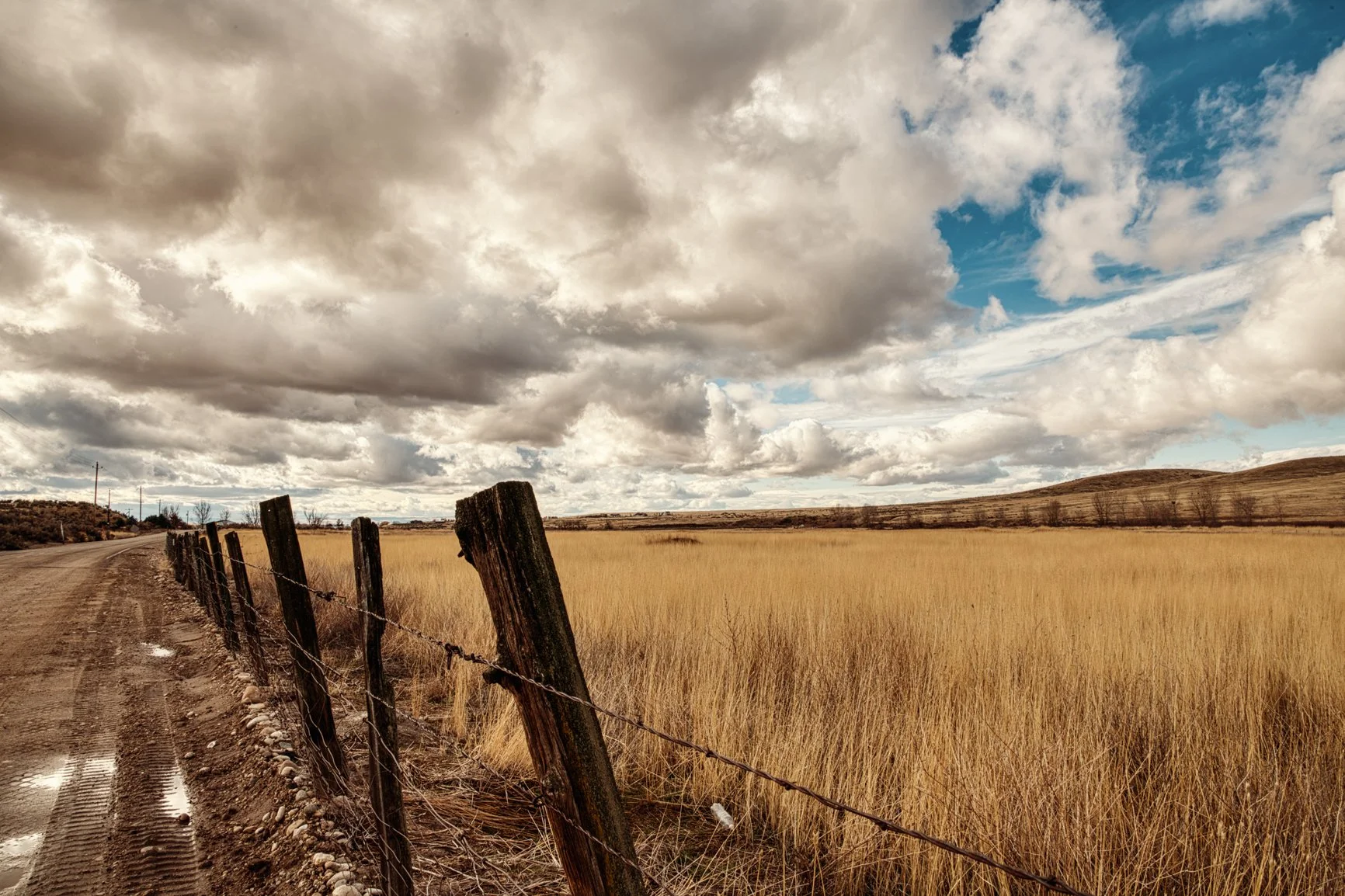 A rugged barbed-wire fence leans against time and weather, guiding the eye across endless golden grasslands toward Idaho’s rolling foothills. Heavy clouds gather above, creating a dramatic sky that contrasts beautifully with the warm tones of late-se