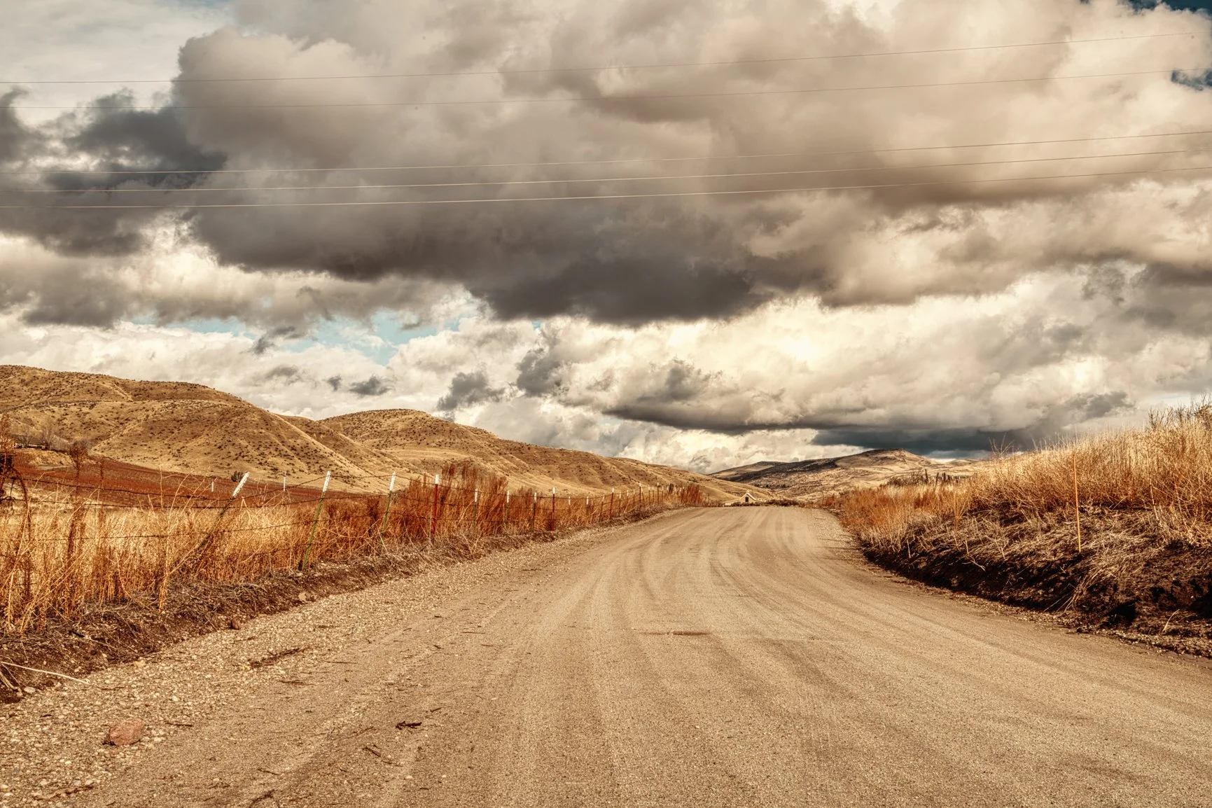 A long dirt road winds through the golden foothills of Idaho beneath a sky heavy with dramatic clouds. The rugged landscape and muted tones echo the quiet solitude of backcountry travel, where every bend hints at discovery and wide-open space stretch