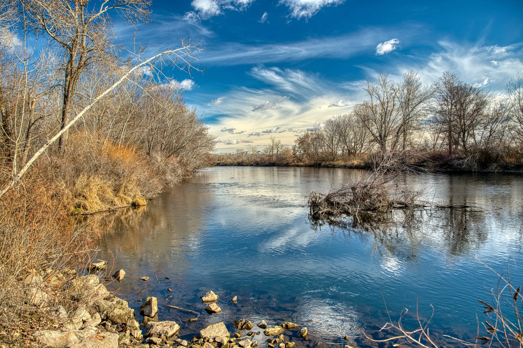 Captured in Star, Idaho, this peaceful Boise River scene reflects the quiet beauty of winter along its winding banks. Bare trees stretch toward a brilliant blue sky as soft clouds drift overhead, mirrored in the river’s gentle flow. The muted tones o