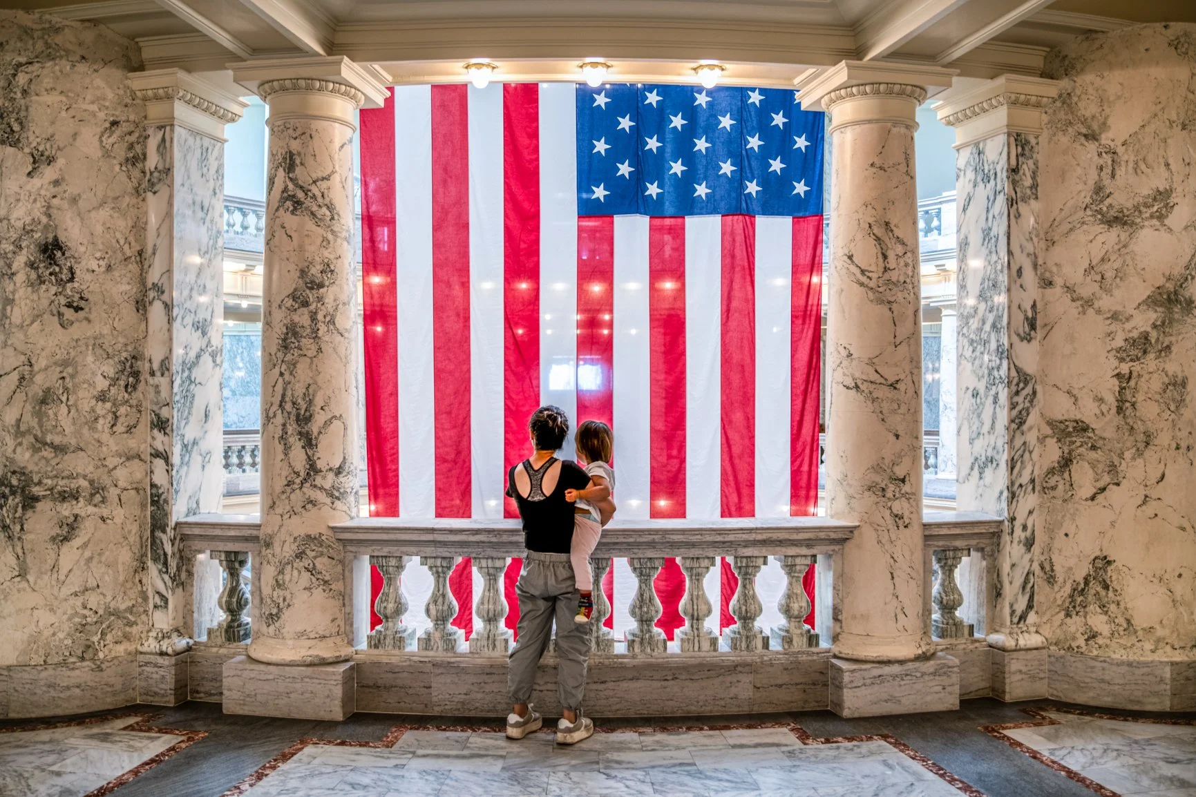 Inside the grand rotunda of the Idaho State Capitol, a mother and child stand quietly before the American flag, sharing a moment of reflection and reverence. Framed by marble columns and soft interior light, the scene speaks to legacy, civic pride, a