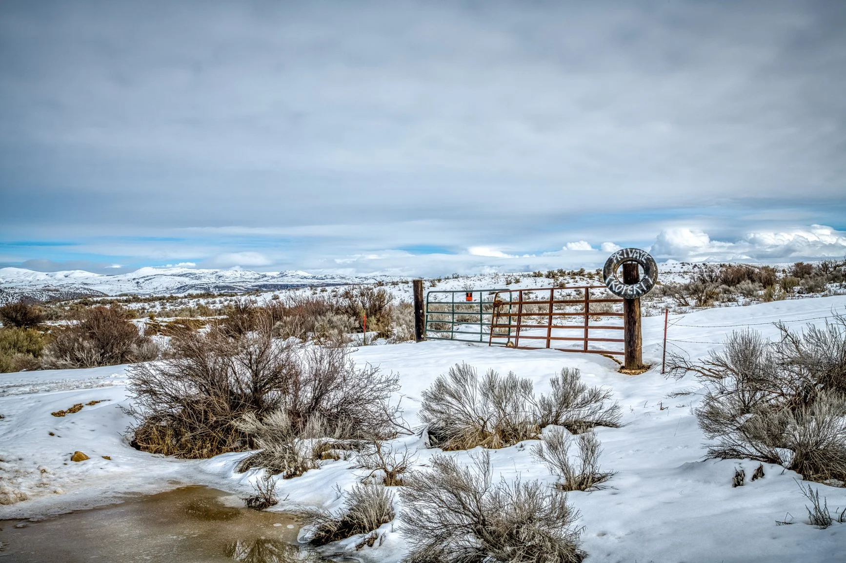Winter settles gently over the Idaho foothills in this serene landscape, where soft snow blankets rolling sagebrush and distant peaks rest beneath a quiet sky. A rustic gate marks the boundary between open country and private land, adding a touch of 