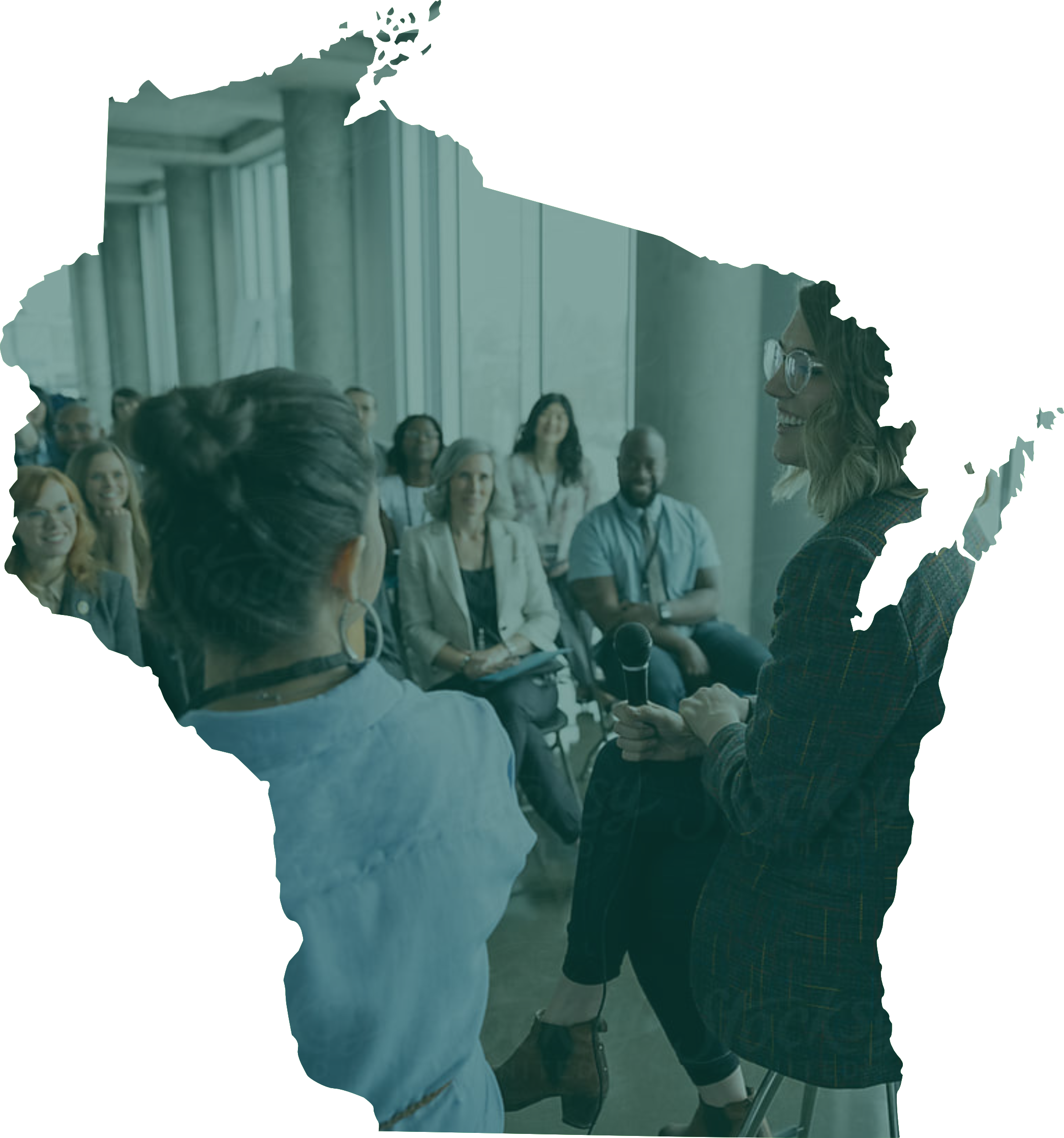 A woman with glasses holding a microphone smiling during a presentation or speech in front of an audience in a modern conference room with large windows and concrete columns.