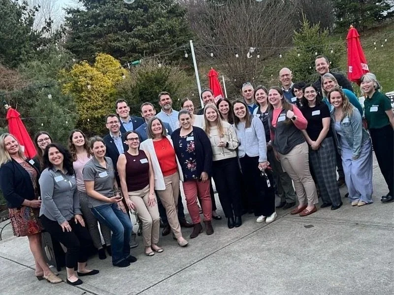 Group of diverse people posing outdoors at a social gathering or event on a concrete patio with red umbrellas and string lights in the background.