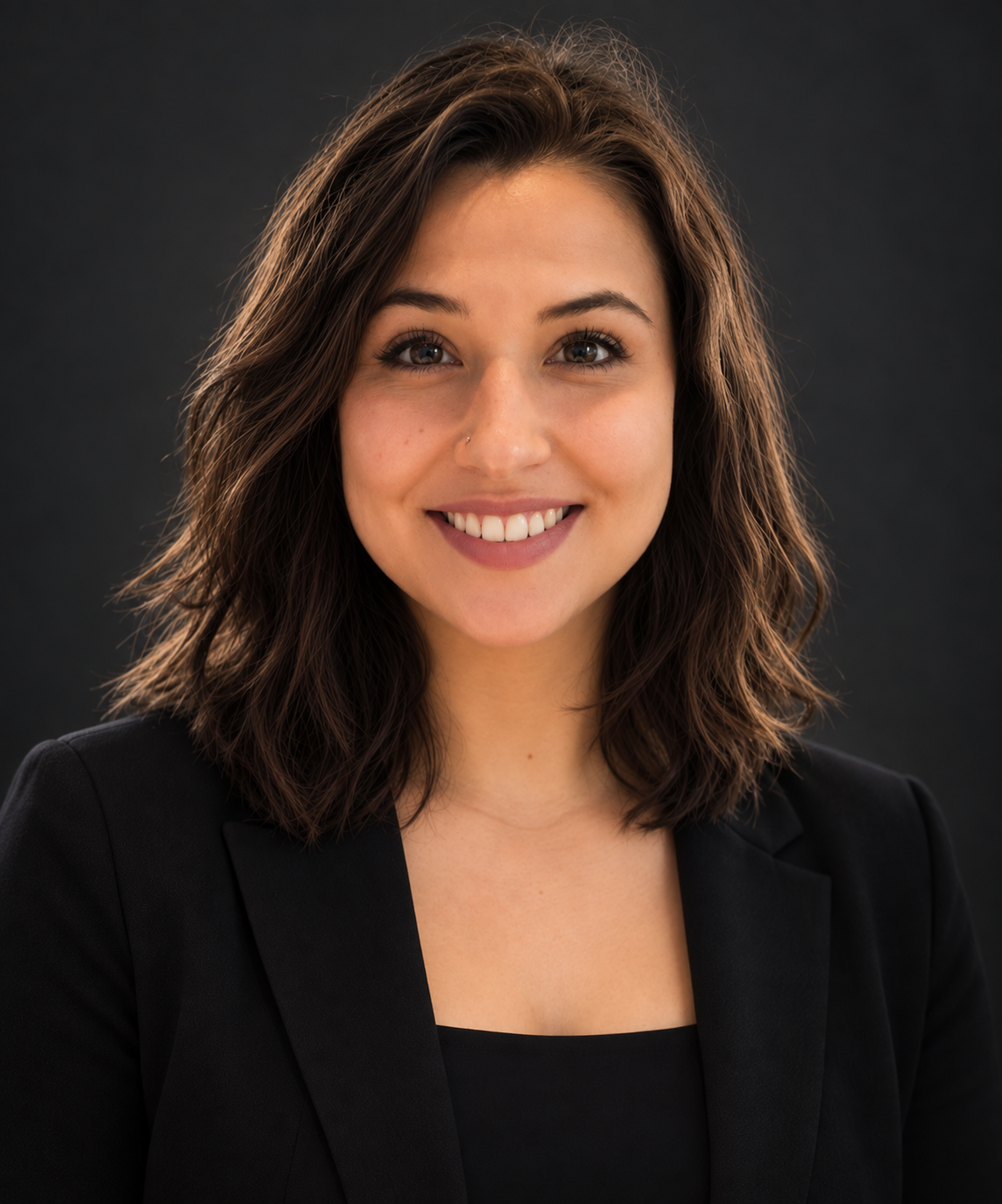 A young woman with shoulder-length brown curly hair, smiling, wearing a black blazer, against a dark background.