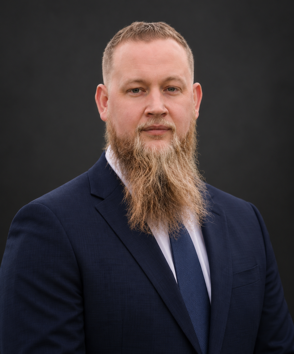 A man with a light beard and mustache wearing a navy blue suit, white shirt, and in front of a dark background.