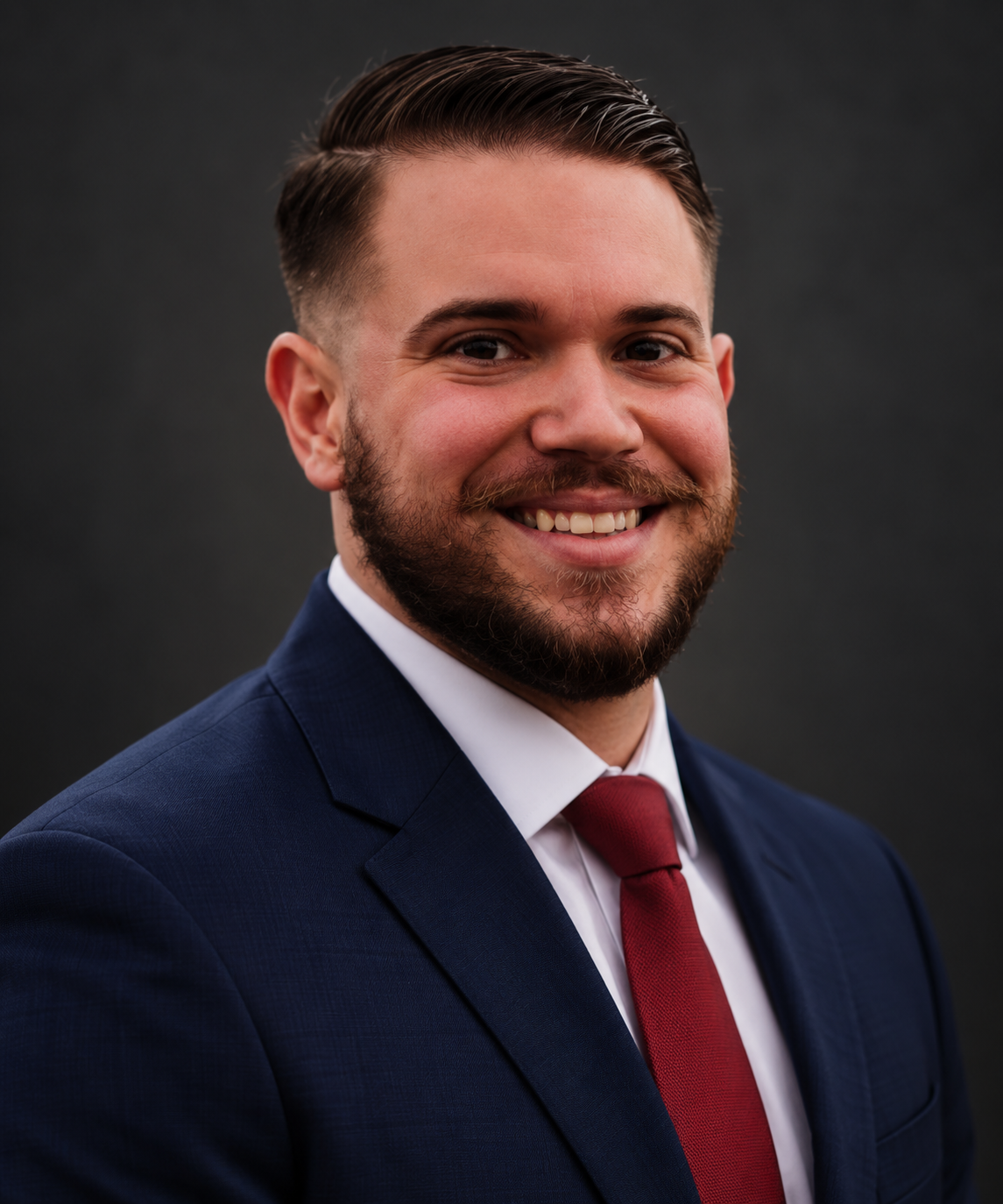 A smiling man with short brown hair and a beard, wearing a navy blue suit, white shirt, and red tie, posing against a dark background.