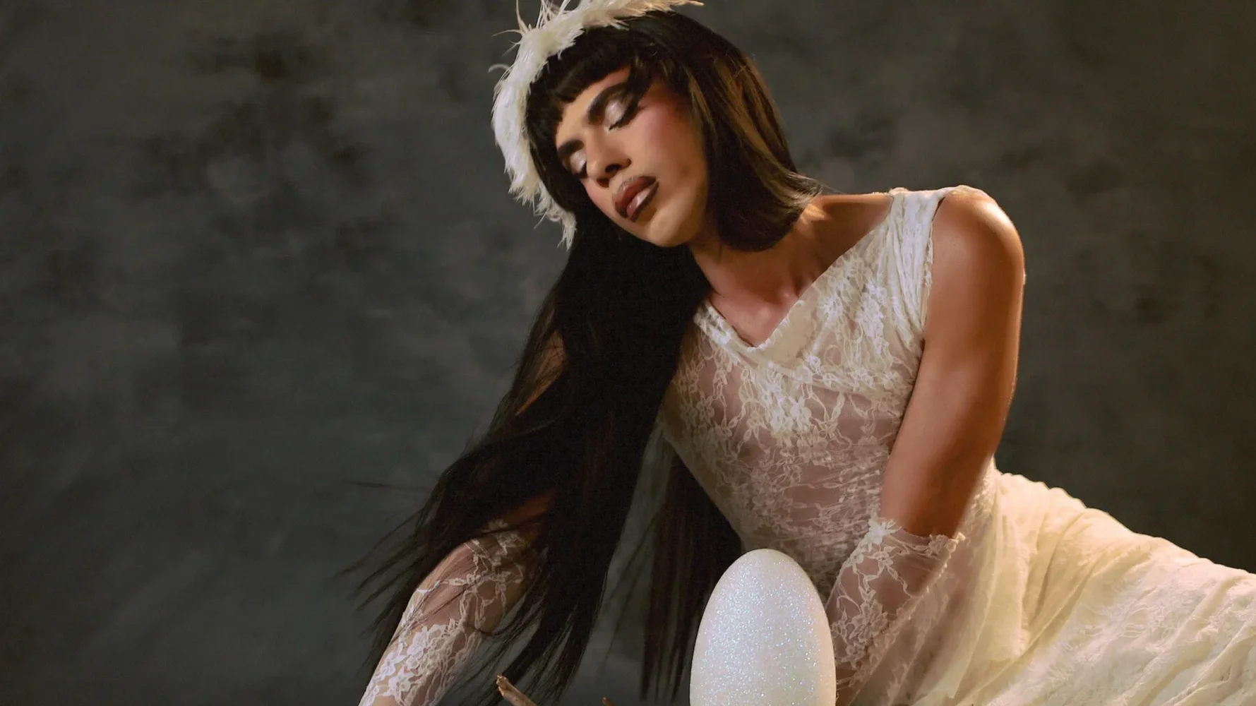 Mujer con vestido de encaje blanco, con ojos cerrados, en una pose expresiva, con fondo oscuro.