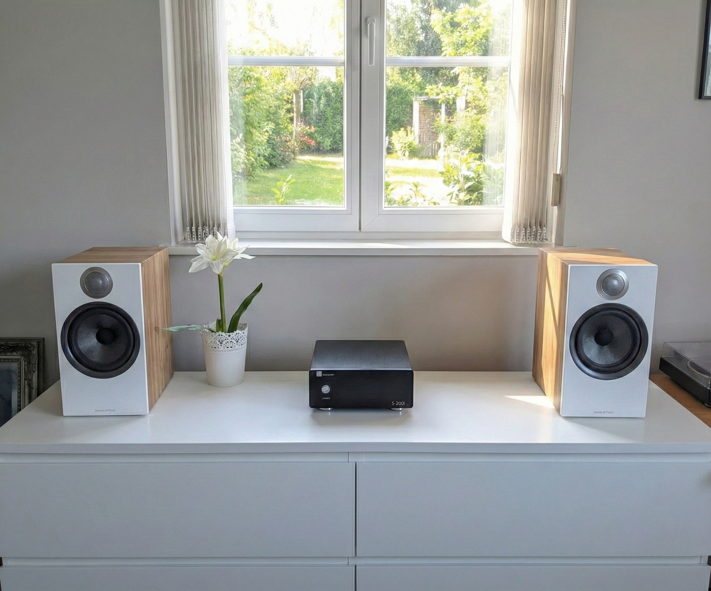 Estante blanca con dos bocinas de madera y una planta con flores blancas, en un hogar con ventana de vista a un jardín verde.