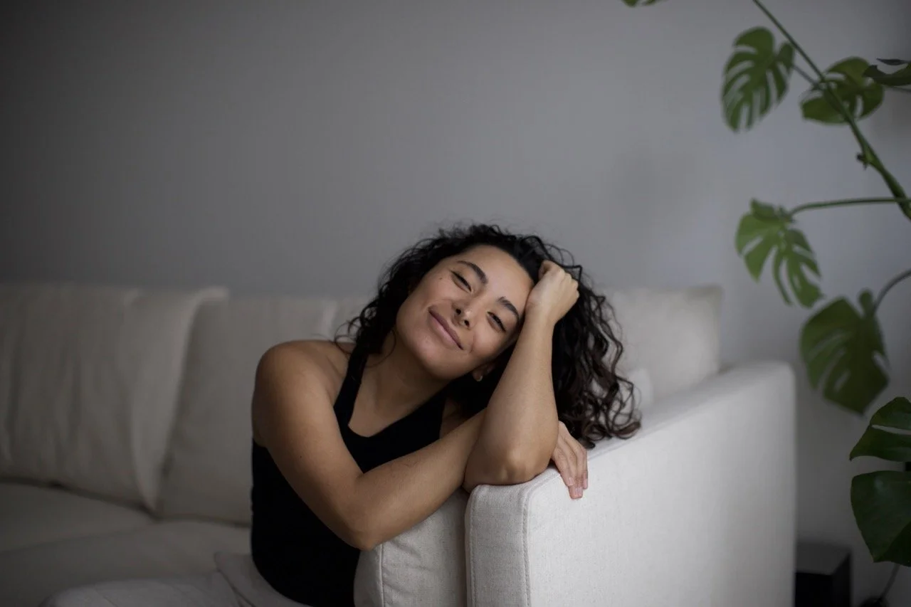 Woman with curly hair smiling while leaning on a white sofa, next to a potted plant.