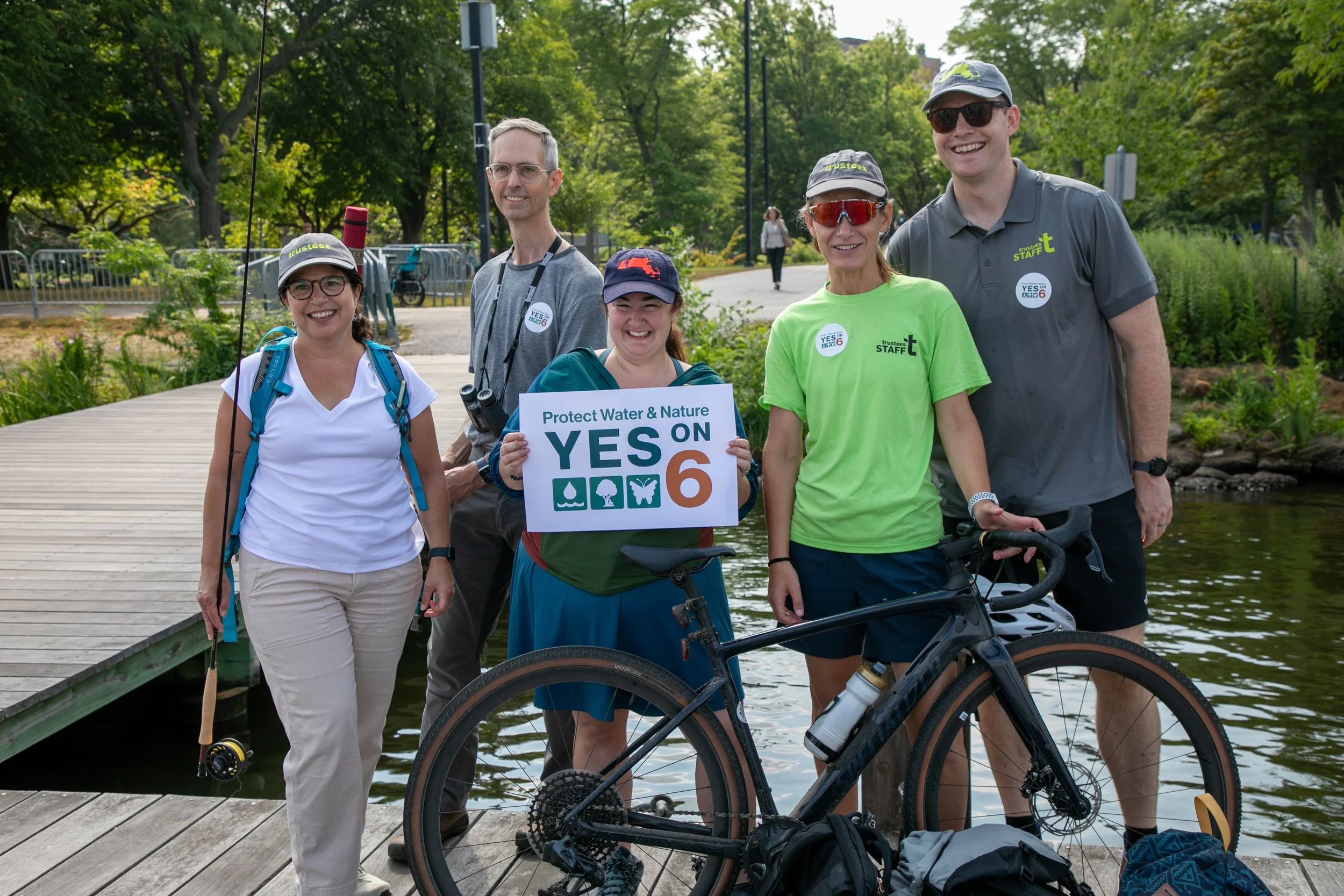 Group of five people standing on a boardwalk near a body of water, smiling at the camera. One person is holding a sign that says 'Protect Water & Nature YES ON 6' and has icons of water, trees, and wildlife. Bicycles are visible in the foreground. The background features green trees and a few pedestrians.