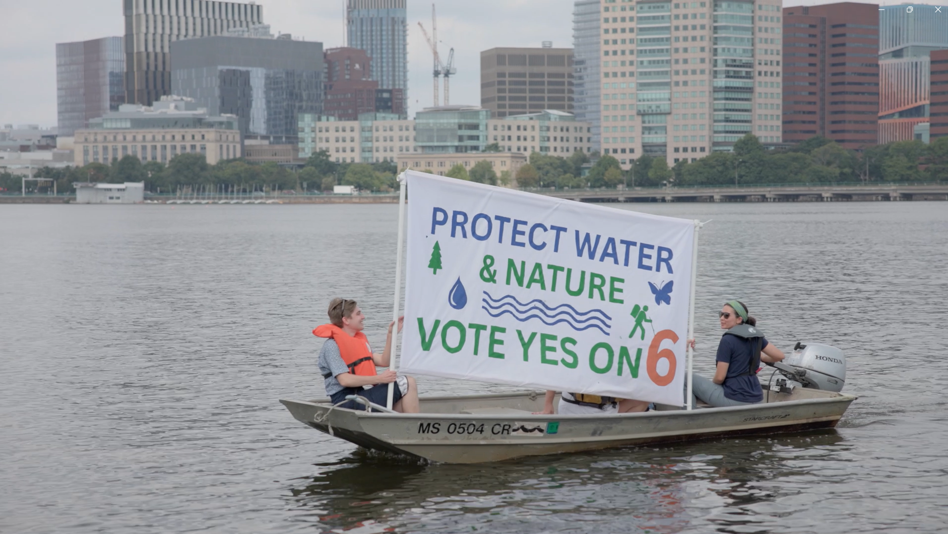 People in a boat holding a sign that reads, "Protect Water & Nature, Vote Yes on 6", with an urban city skyline in the background.