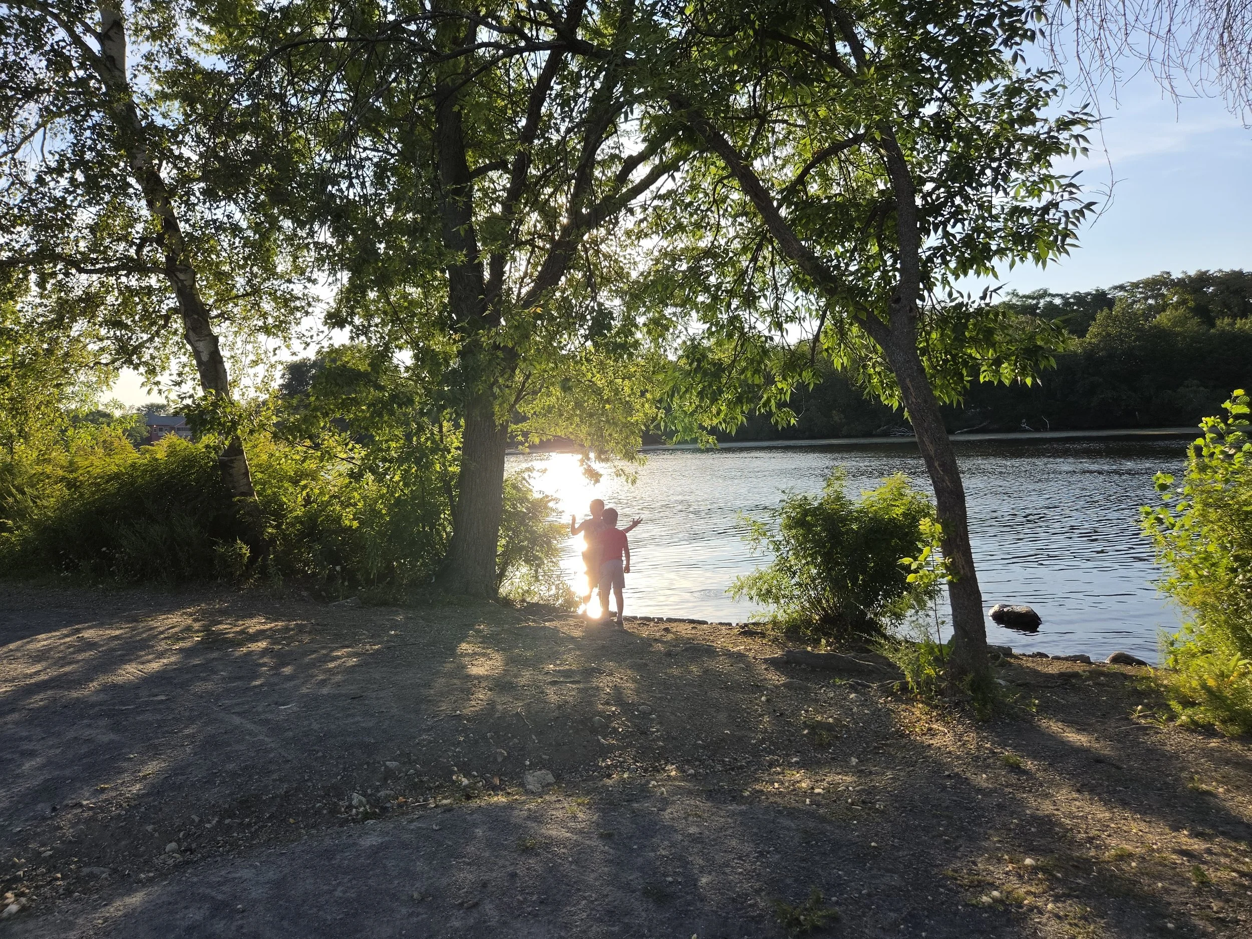 A person standing at the edge of a river during sunset with trees and clear sky in the background.