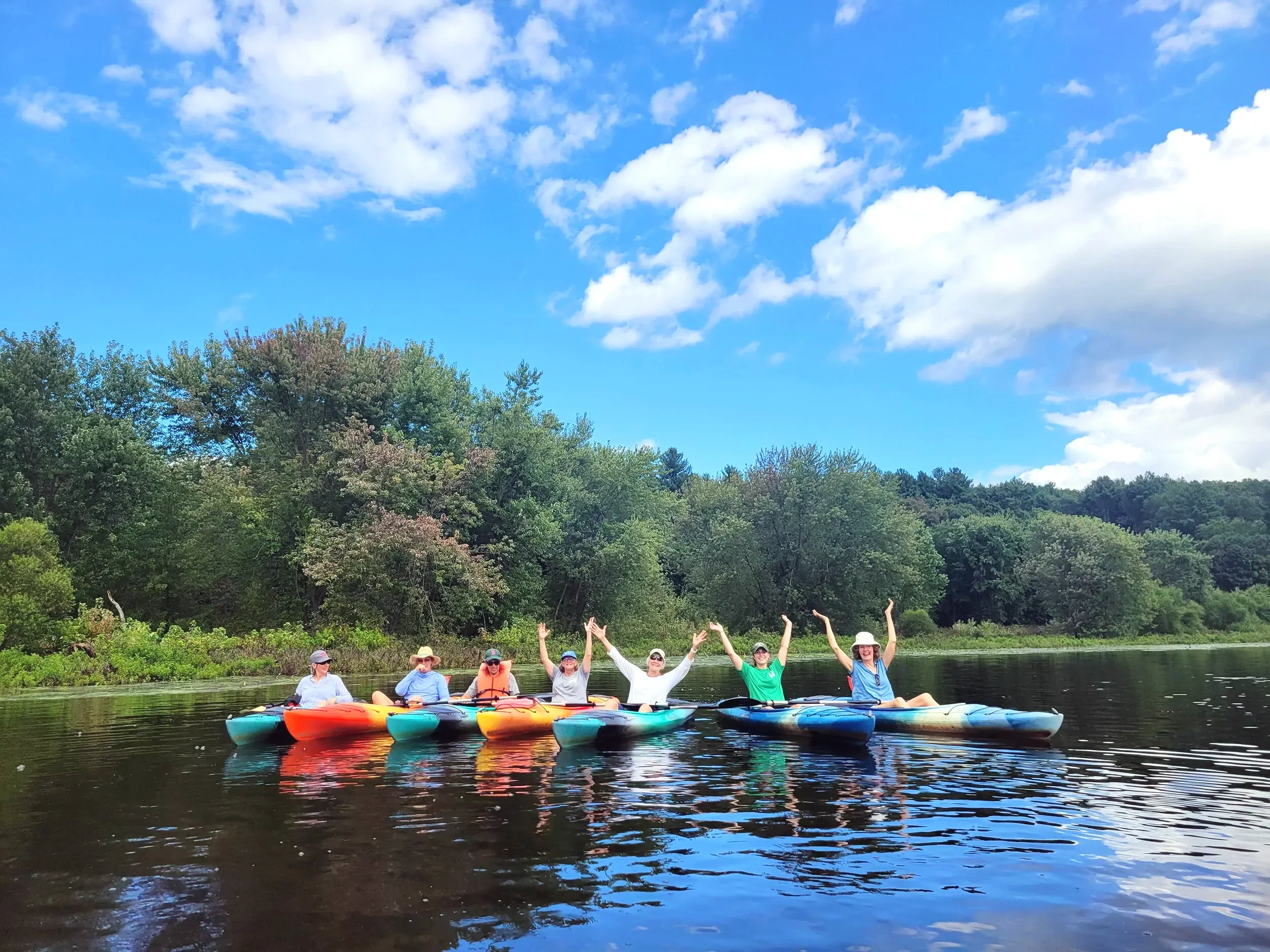 Group of people kayaking on a calm river, surrounded by green trees and a bright blue sky with clouds.